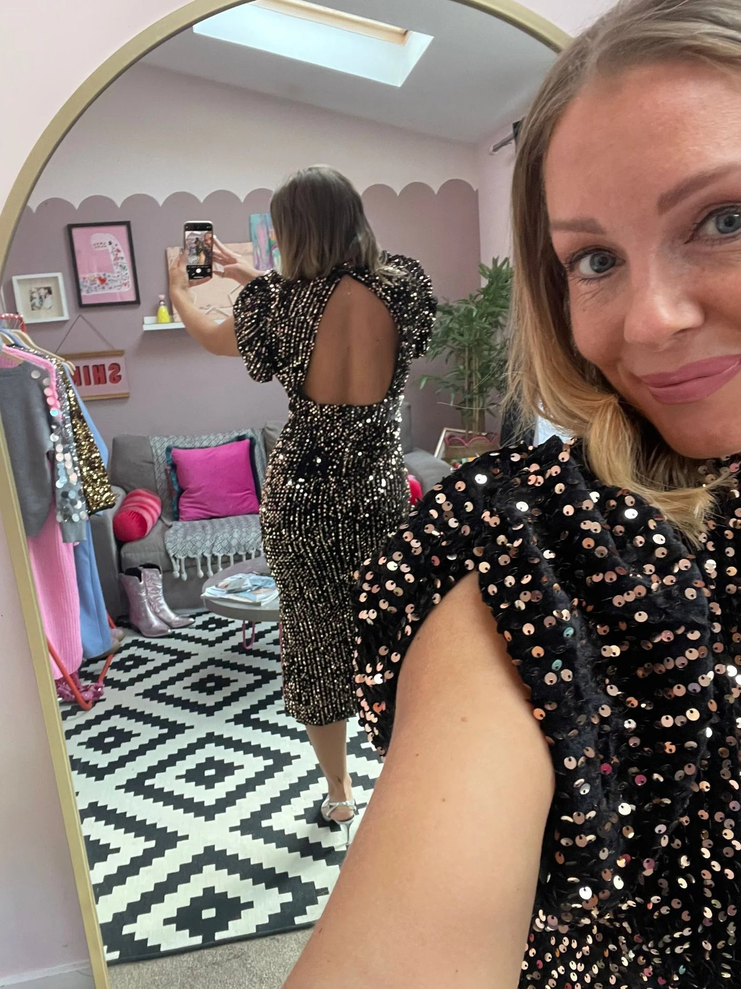Woman taking a mirror selfie wearing a black sequin dress in a room with a patterned rug and furniture.