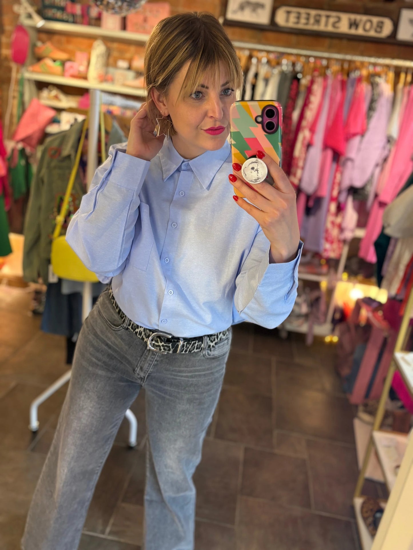 Woman taking a mirror selfie in a clothing store wearing a light blue shirt and jeans.