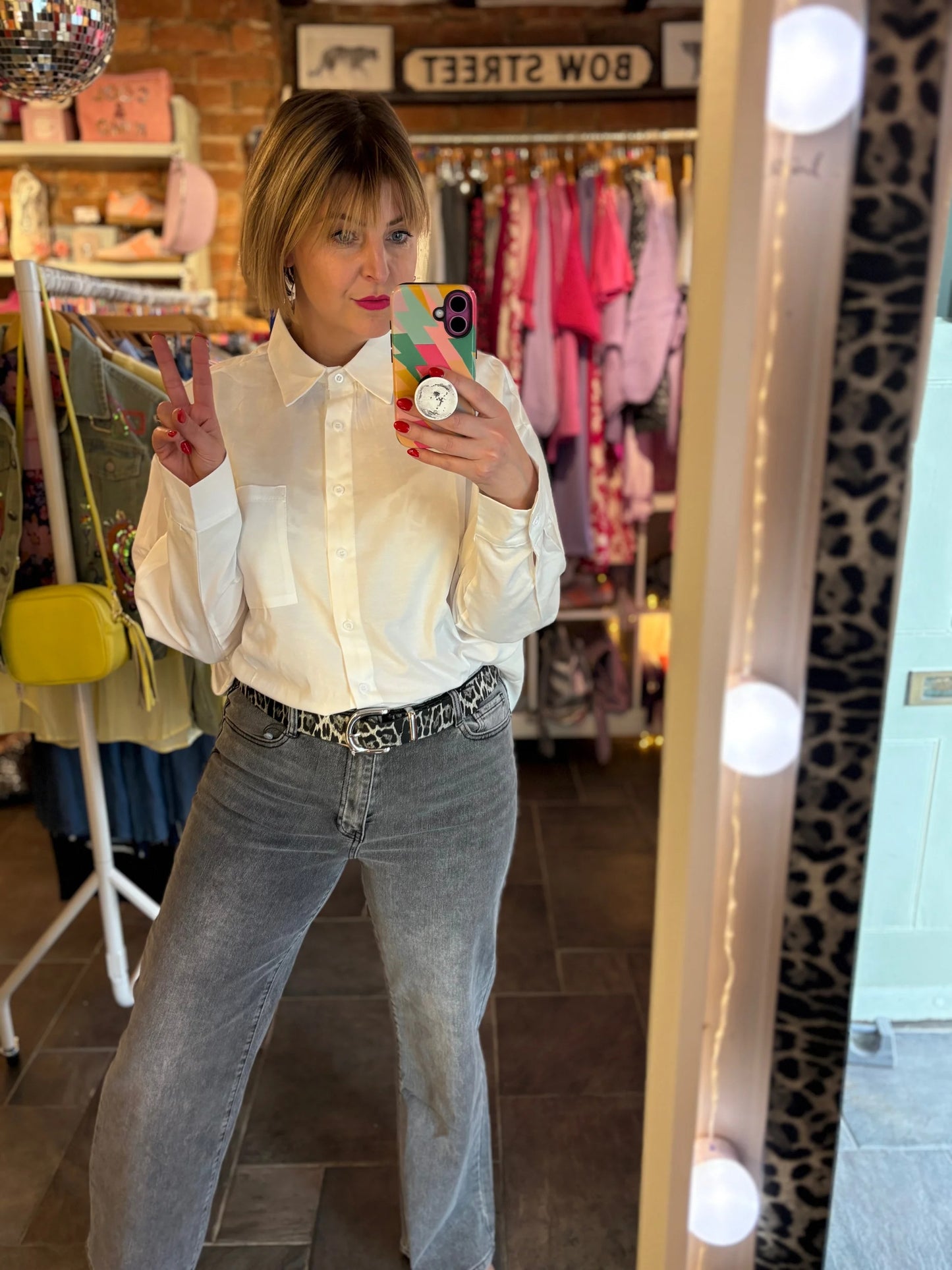 Person taking a mirror selfie wearing a white shirt and gray jeans in a store.
