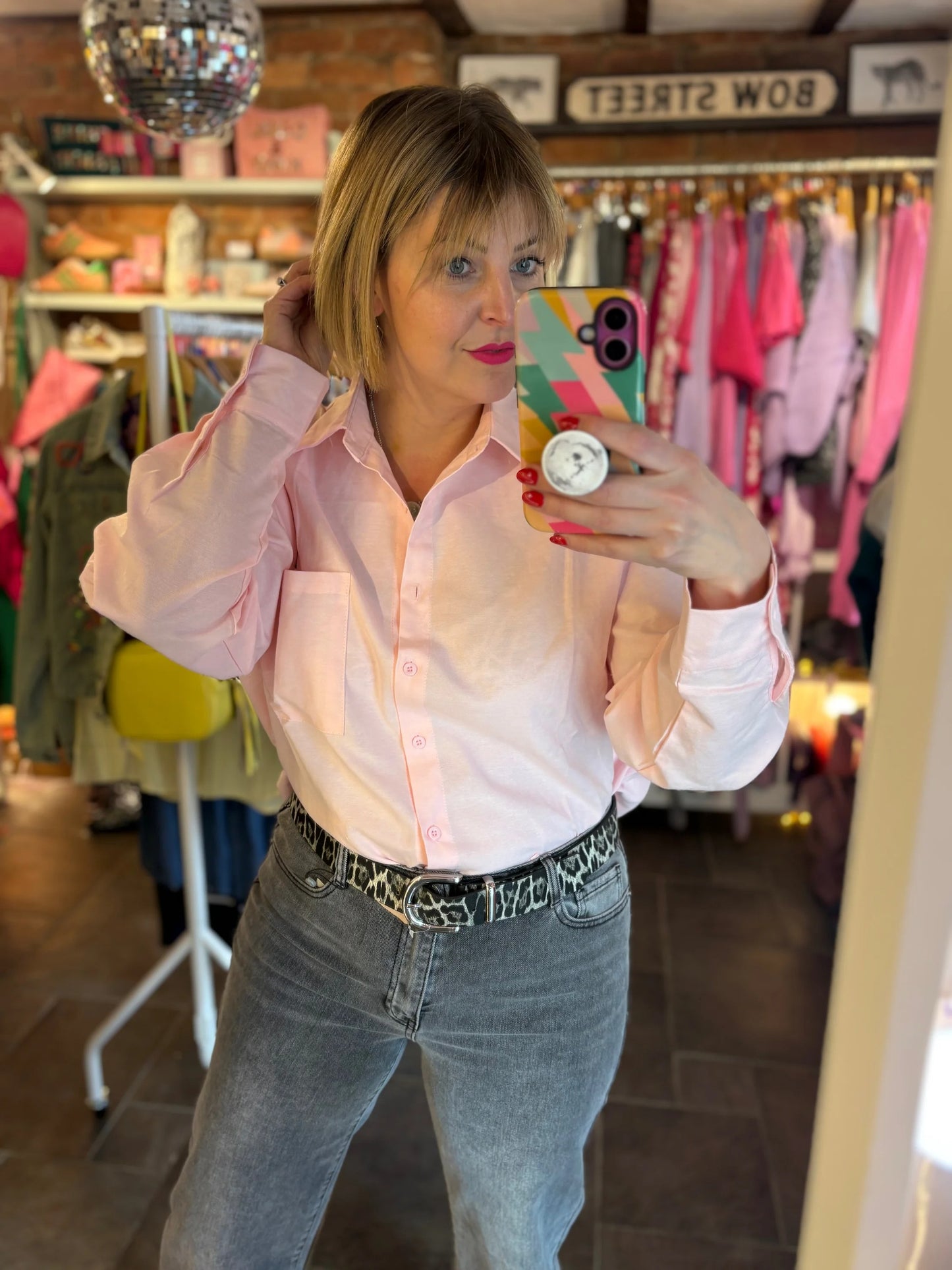 Woman taking a mirror selfie in a clothing store wearing a pink shirt and jeans.