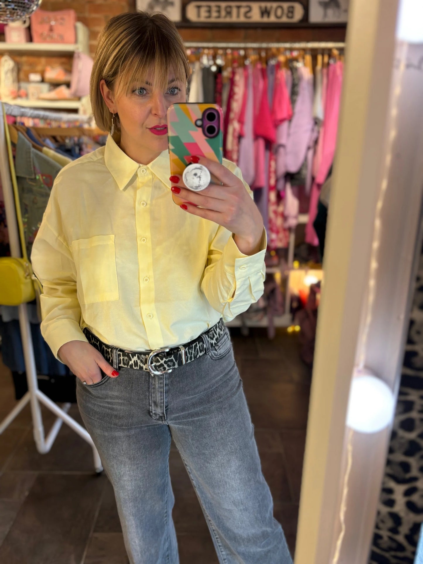 Person taking a mirror selfie wearing a yellow shirt and blue jeans in a clothing store.