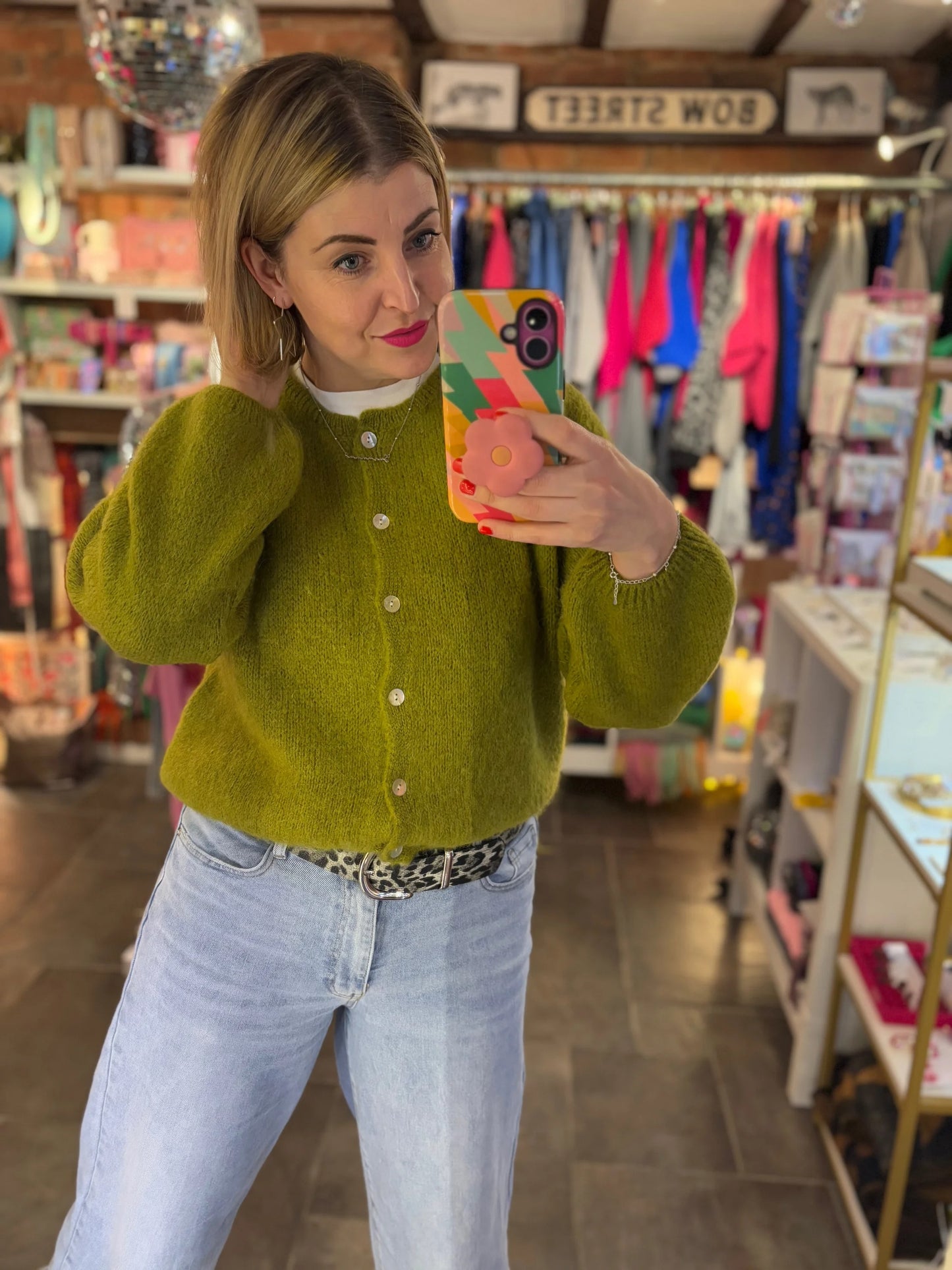 Person taking a mirror selfie wearing a green cardigan and blue jeans in a store.