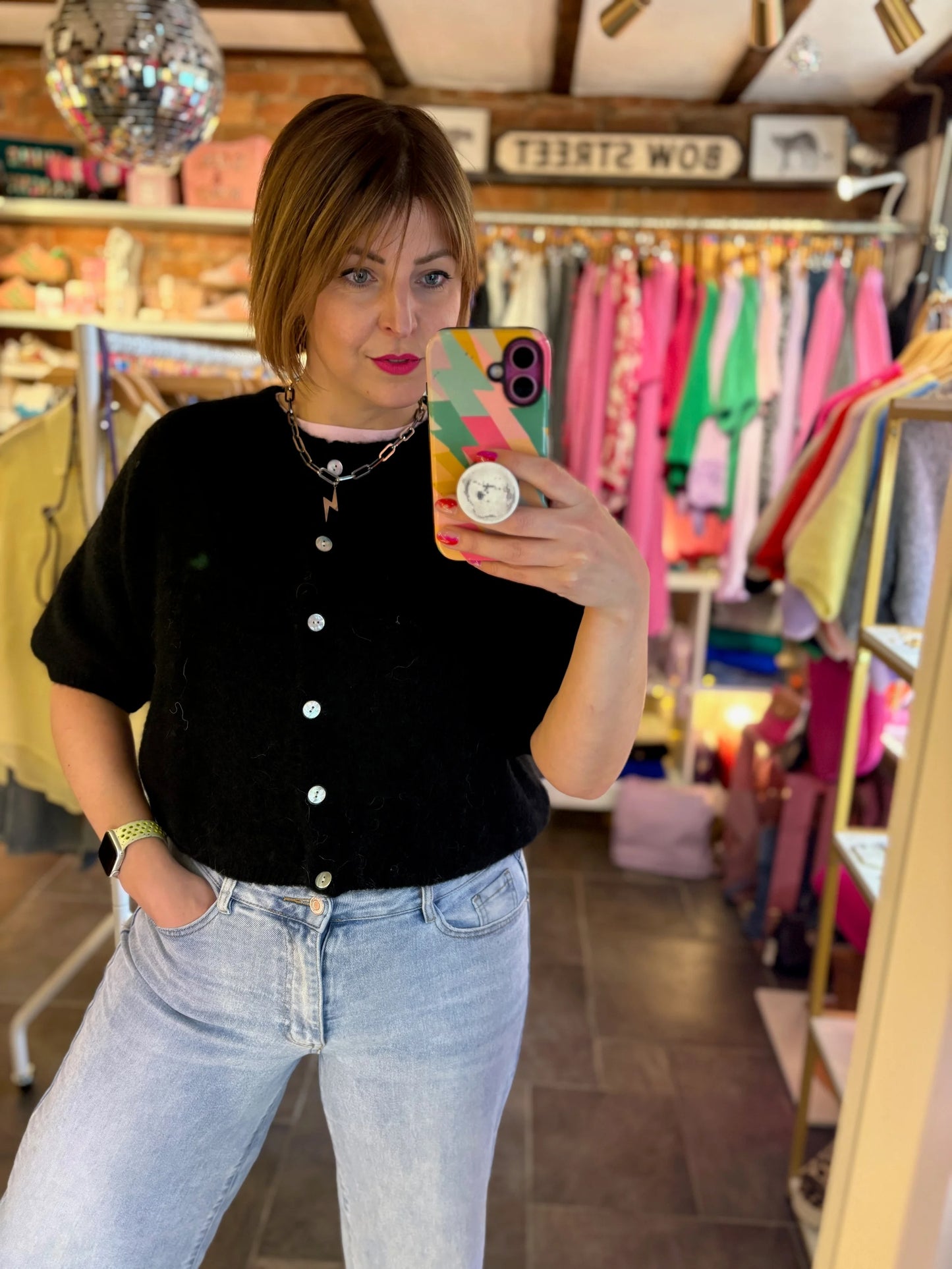 Woman taking a mirror selfie in a clothing store with colorful racks in the background.