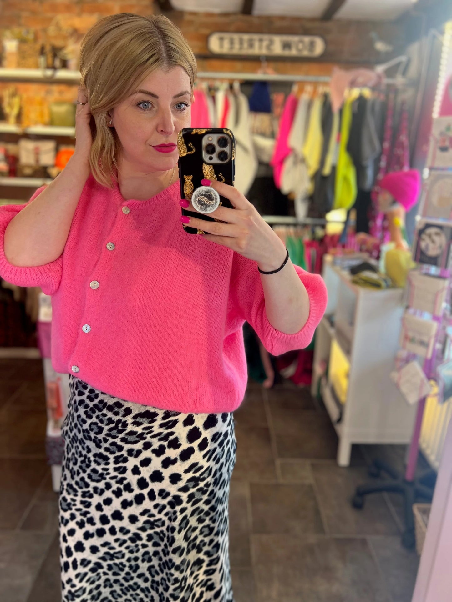 Woman taking a mirror selfie wearing a bright pink cardigan and leopard print skirt in a store.