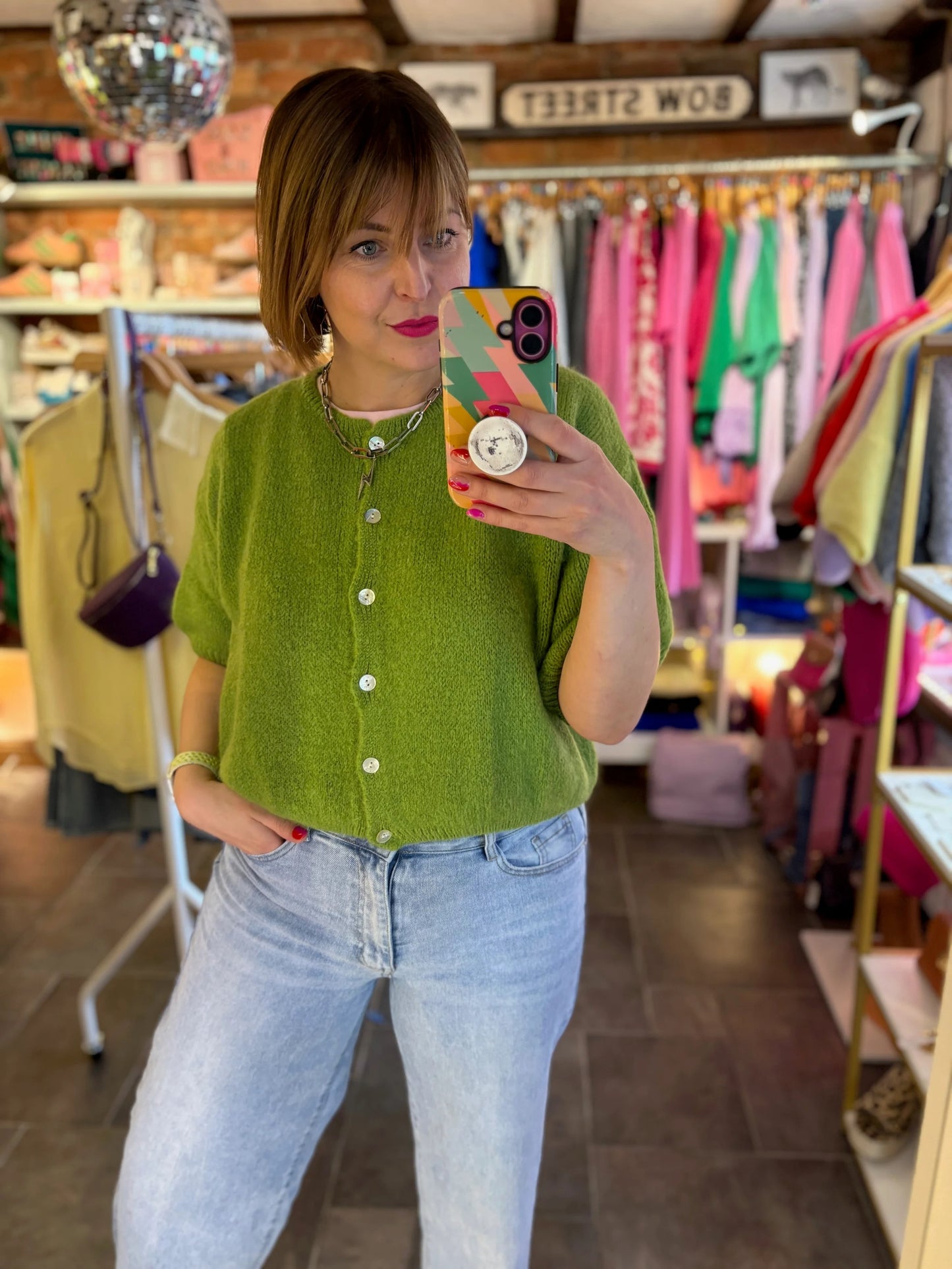 Person wearing a green shirt and blue jeans taking a mirror selfie in a clothing store.