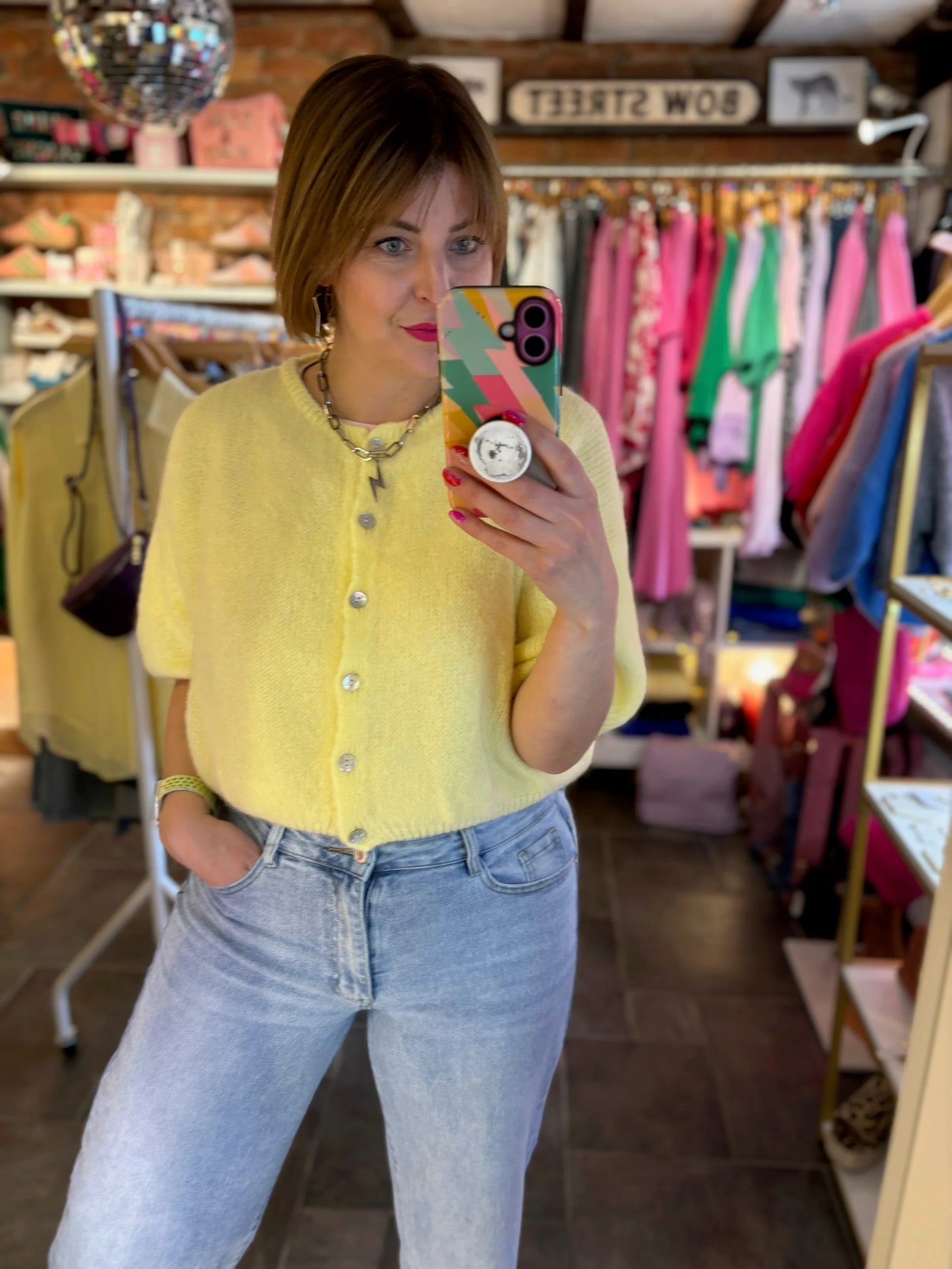 Woman taking a mirror selfie wearing a yellow shirt and blue jeans in a clothing store.
