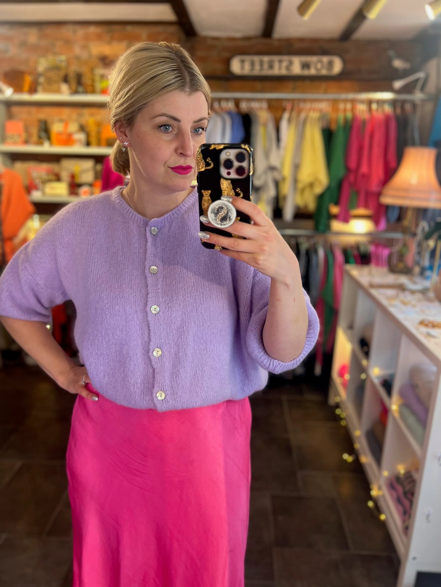 Woman taking a mirror selfie wearing a purple cardigan and pink skirt in a store.