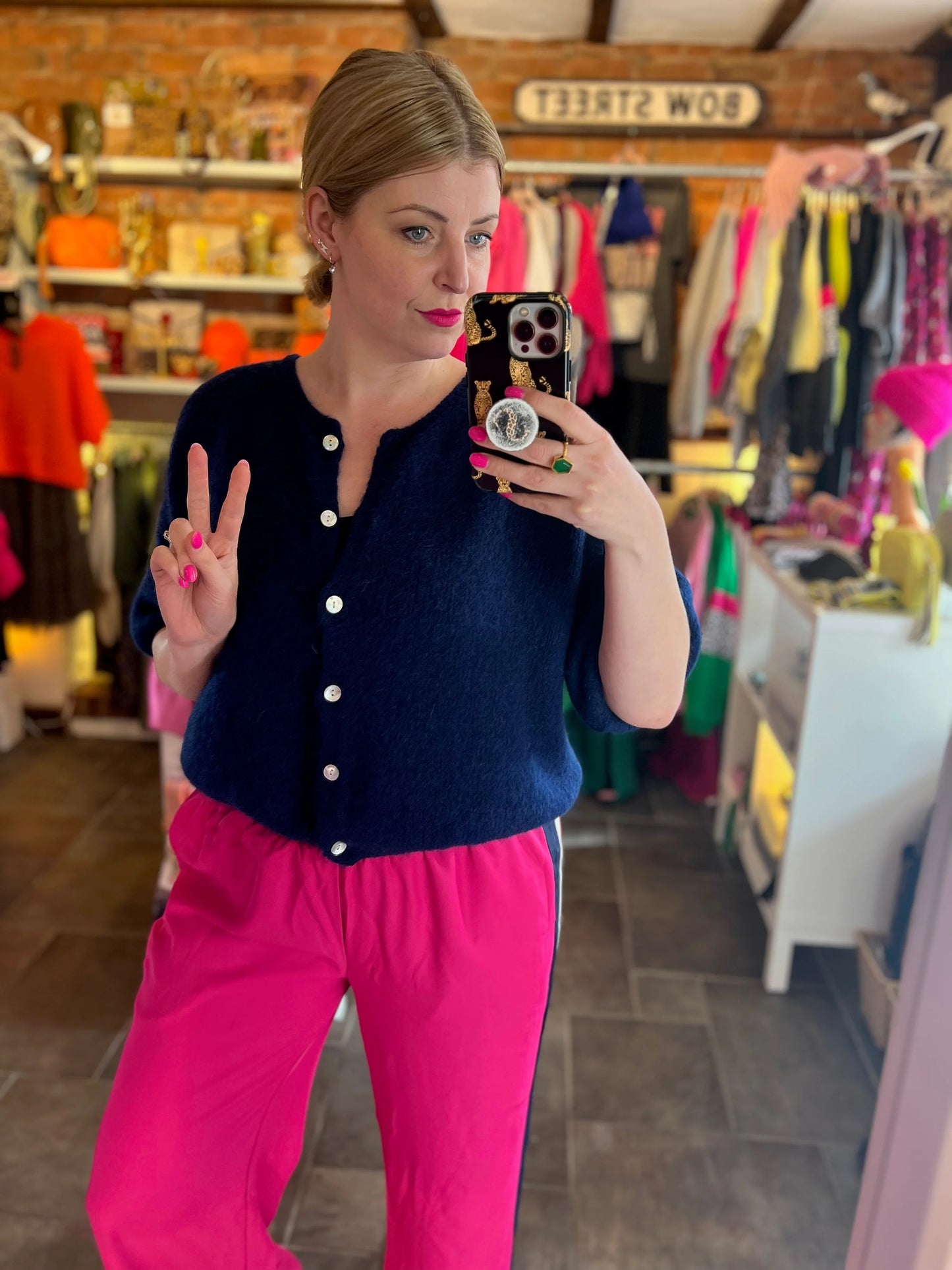 Woman taking a mirror selfie in a clothing store wearing a navy cardigan and pink pants.