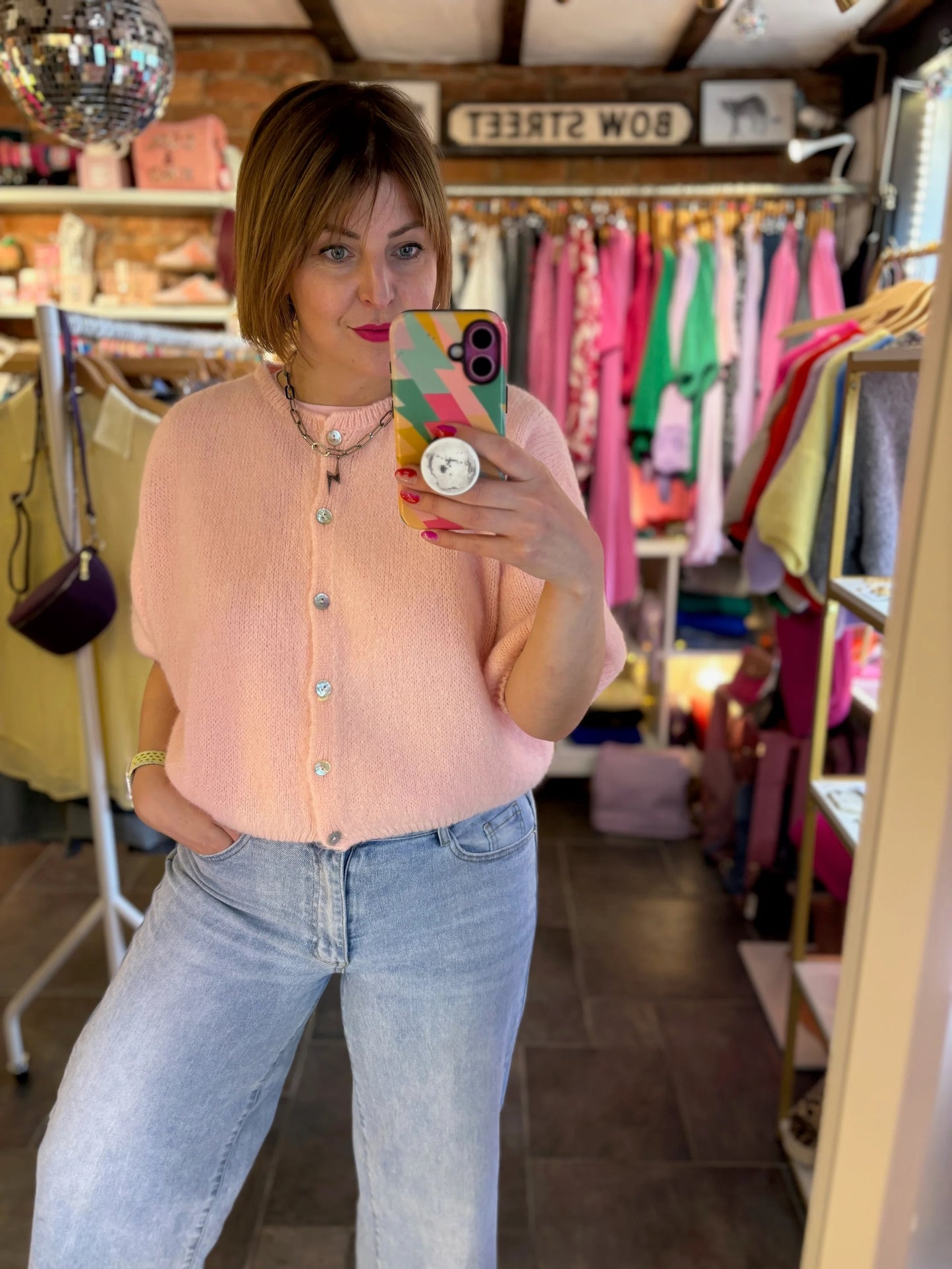 Woman taking a mirror selfie wearing a pink cardigan and blue jeans in a clothing store.