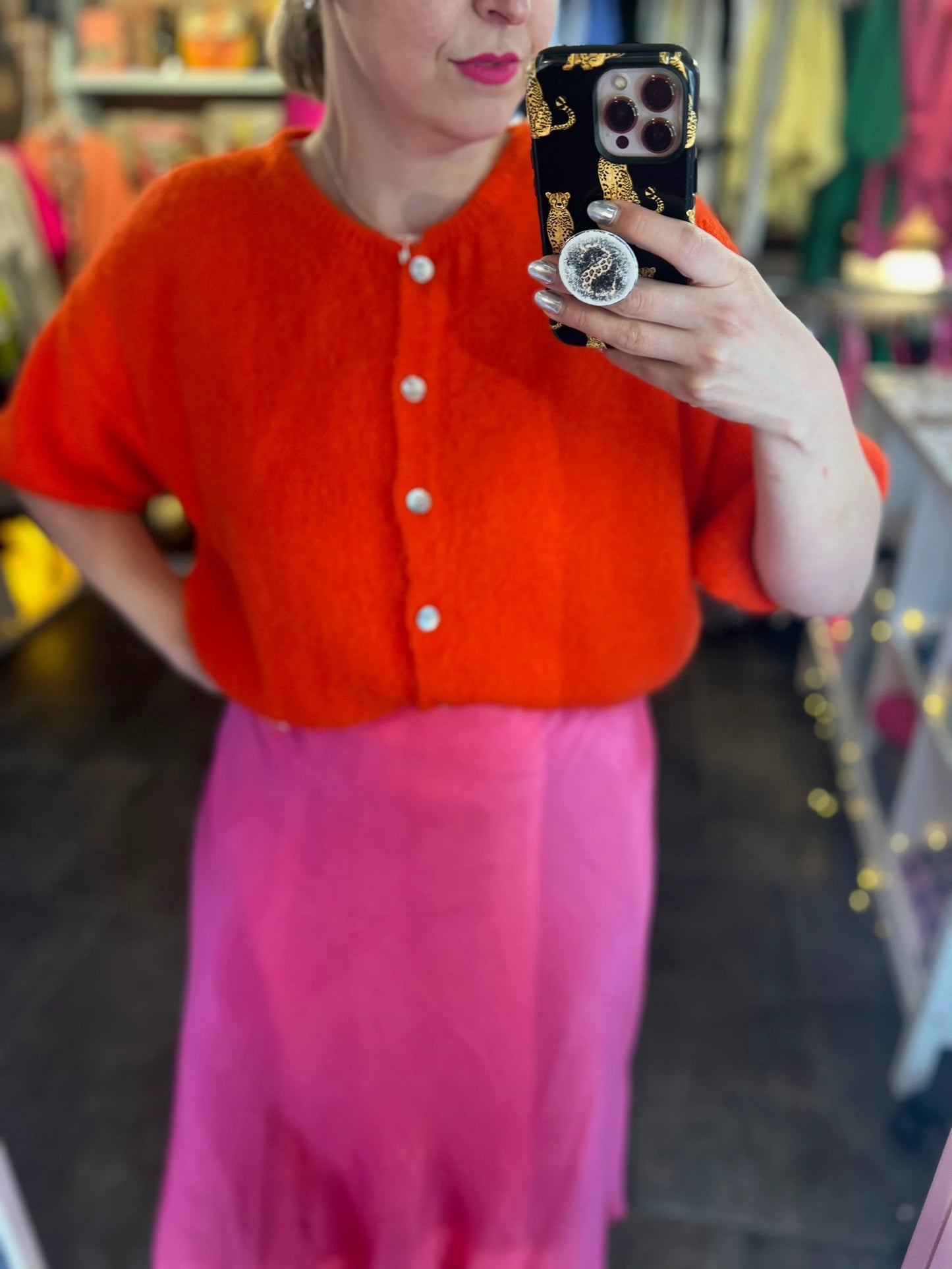 Person wearing an orange cardigan and pink skirt taking a mirror selfie.