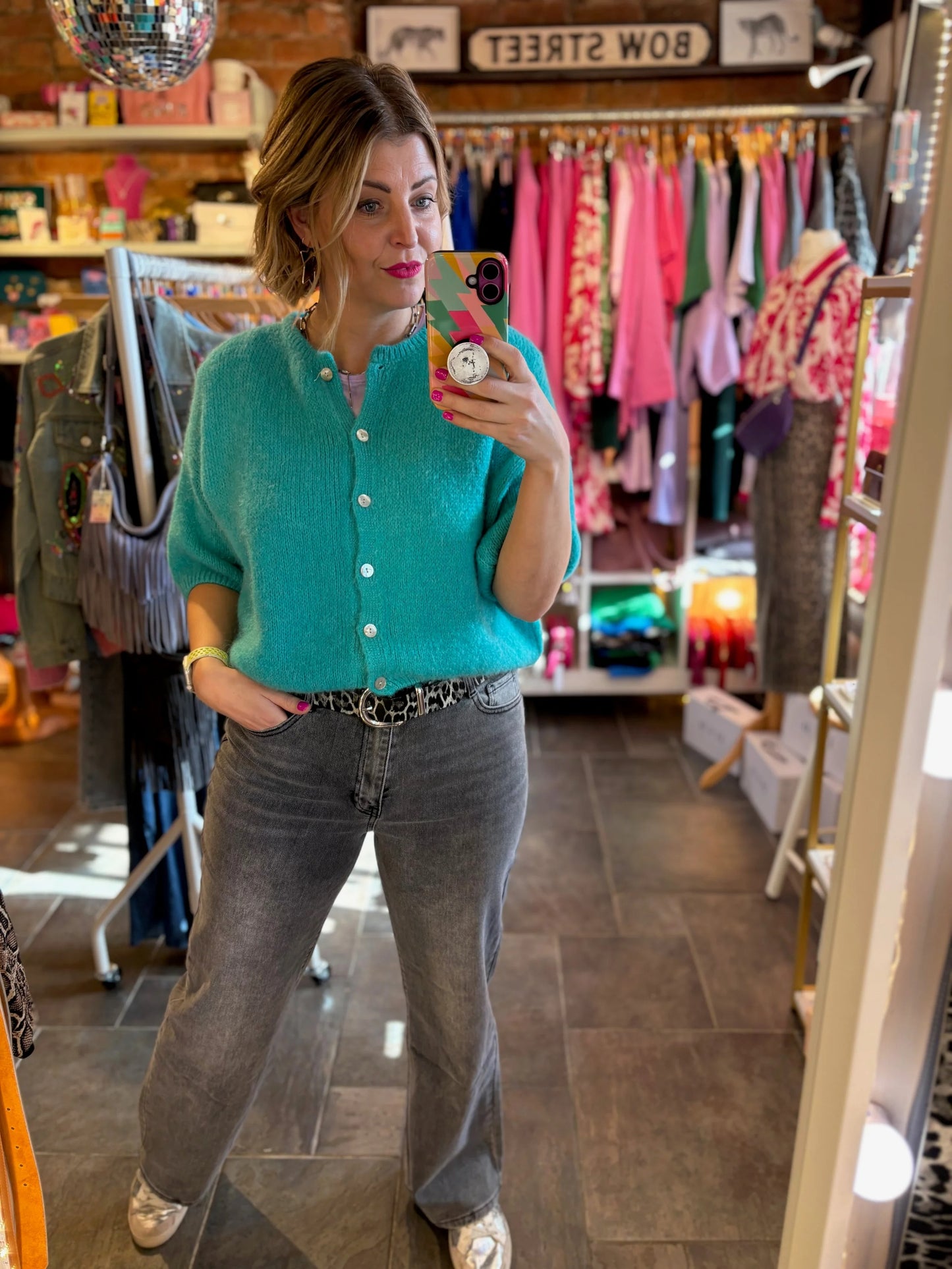 Woman taking a mirror selfie wearing a teal cardigan and gray jeans in a clothing store.