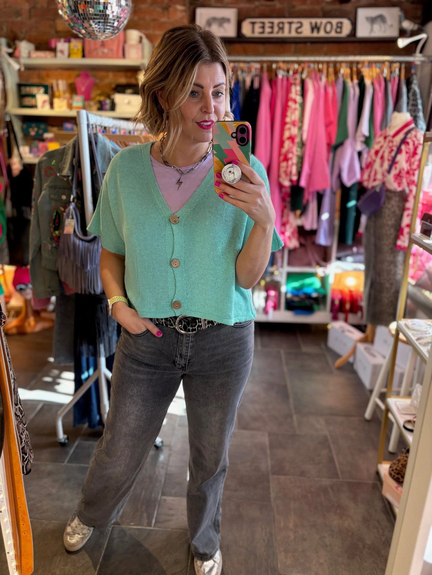 Woman taking a mirror selfie in a clothing store wearing a green cardigan and gray jeans.