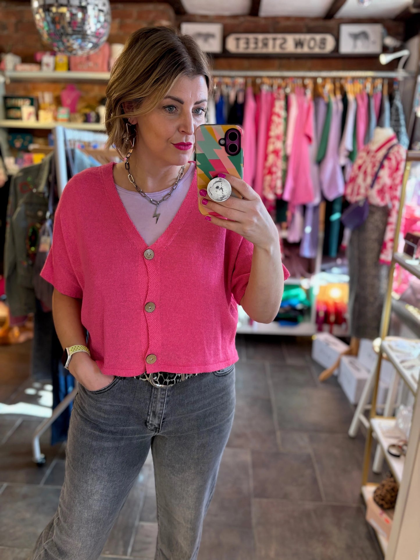Woman taking a mirror selfie wearing a pink cardigan and gray jeans in a clothing store.