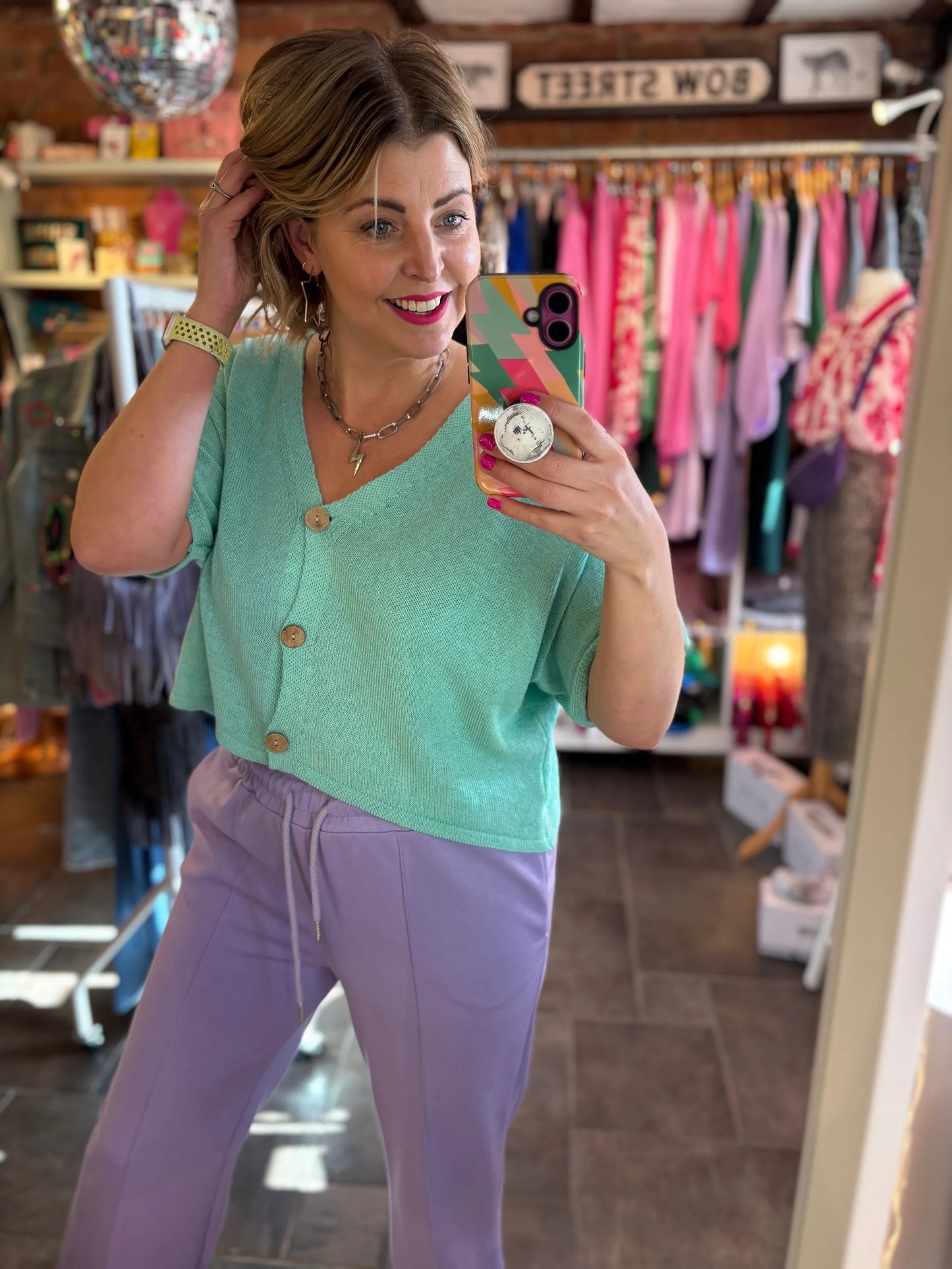 Woman taking a mirror selfie wearing a green cardigan and purple pants in a store.
