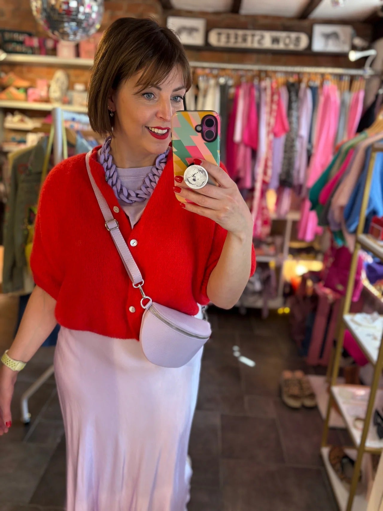 Woman taking a mirror selfie in a clothing store wearing a red cardigan and white skirt.