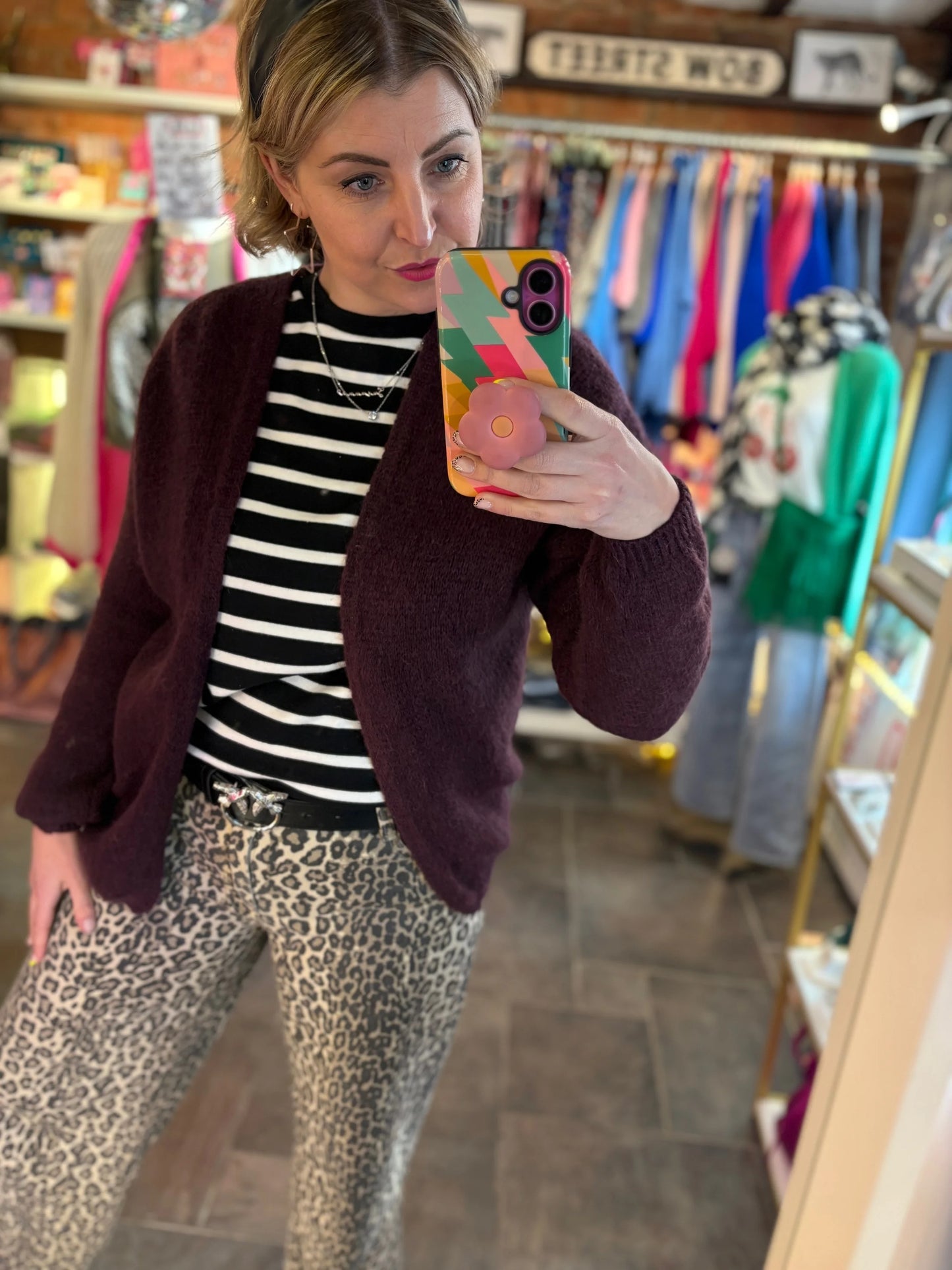 Woman taking a mirror selfie in a store wearing a striped shirt, maroon cardigan, and leopard print pants.