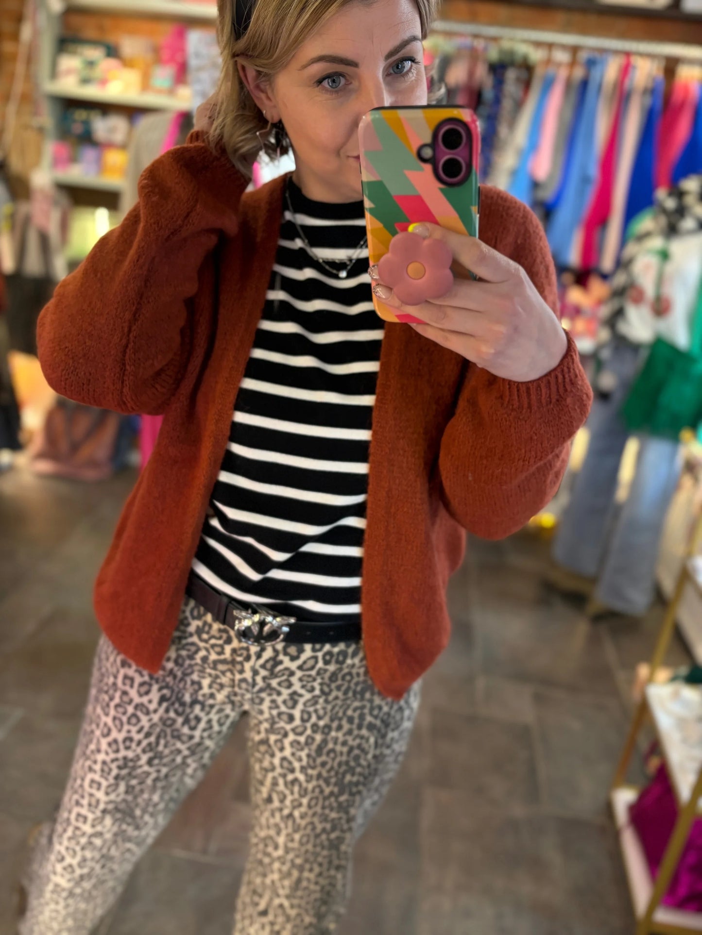 Person taking a mirror selfie wearing a rust-colored cardigan, black and white striped shirt, and leopard print pants in a store.