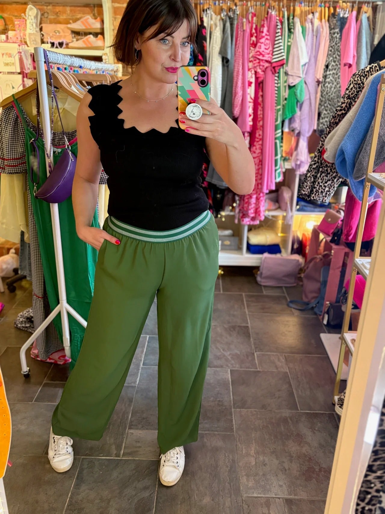 Woman taking a mirror selfie wearing a black top and green pants in a clothing store.