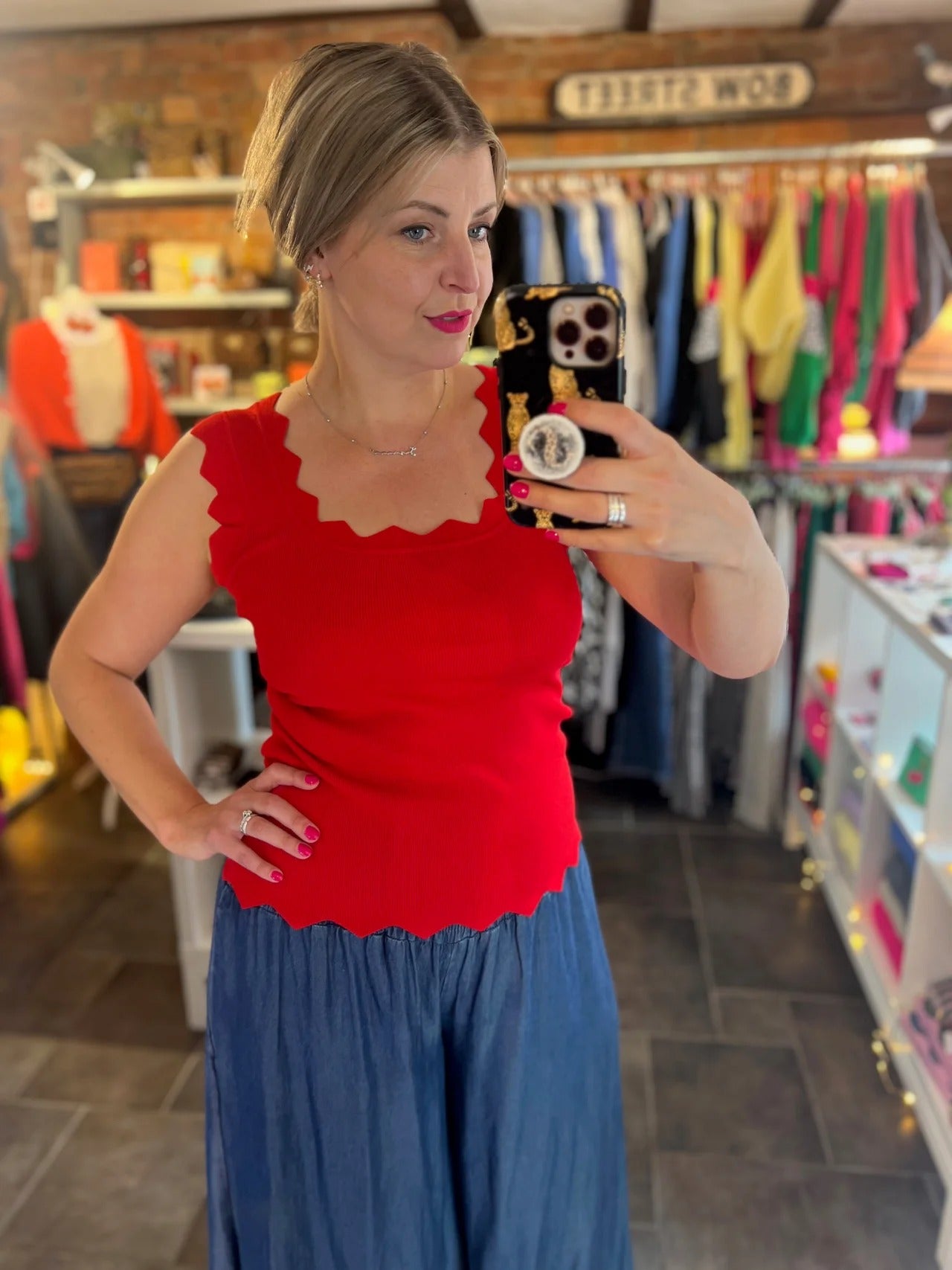 Woman taking a mirror selfie wearing a red top and blue pants in a store.