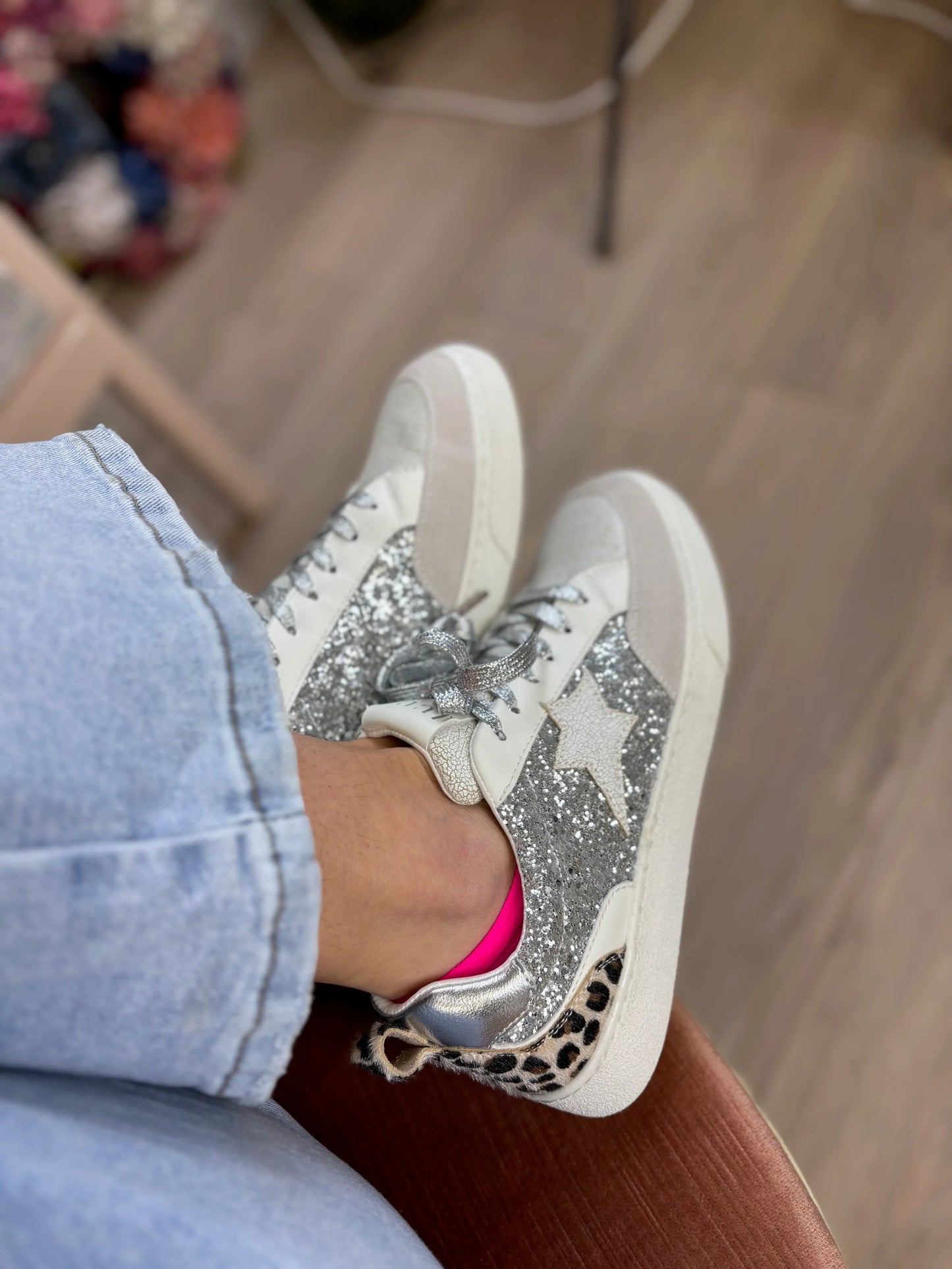 White sneakers with glittery design worn by a person sitting on a wooden floor.