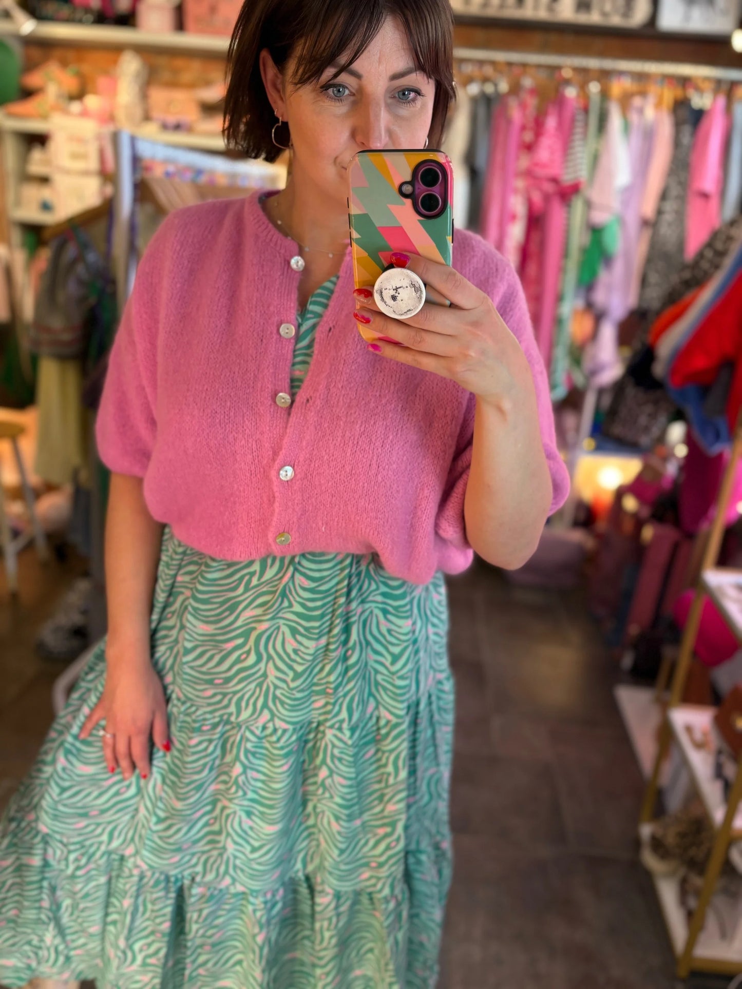 Person taking a mirror selfie wearing a pink cardigan and green patterned dress in a clothing store.