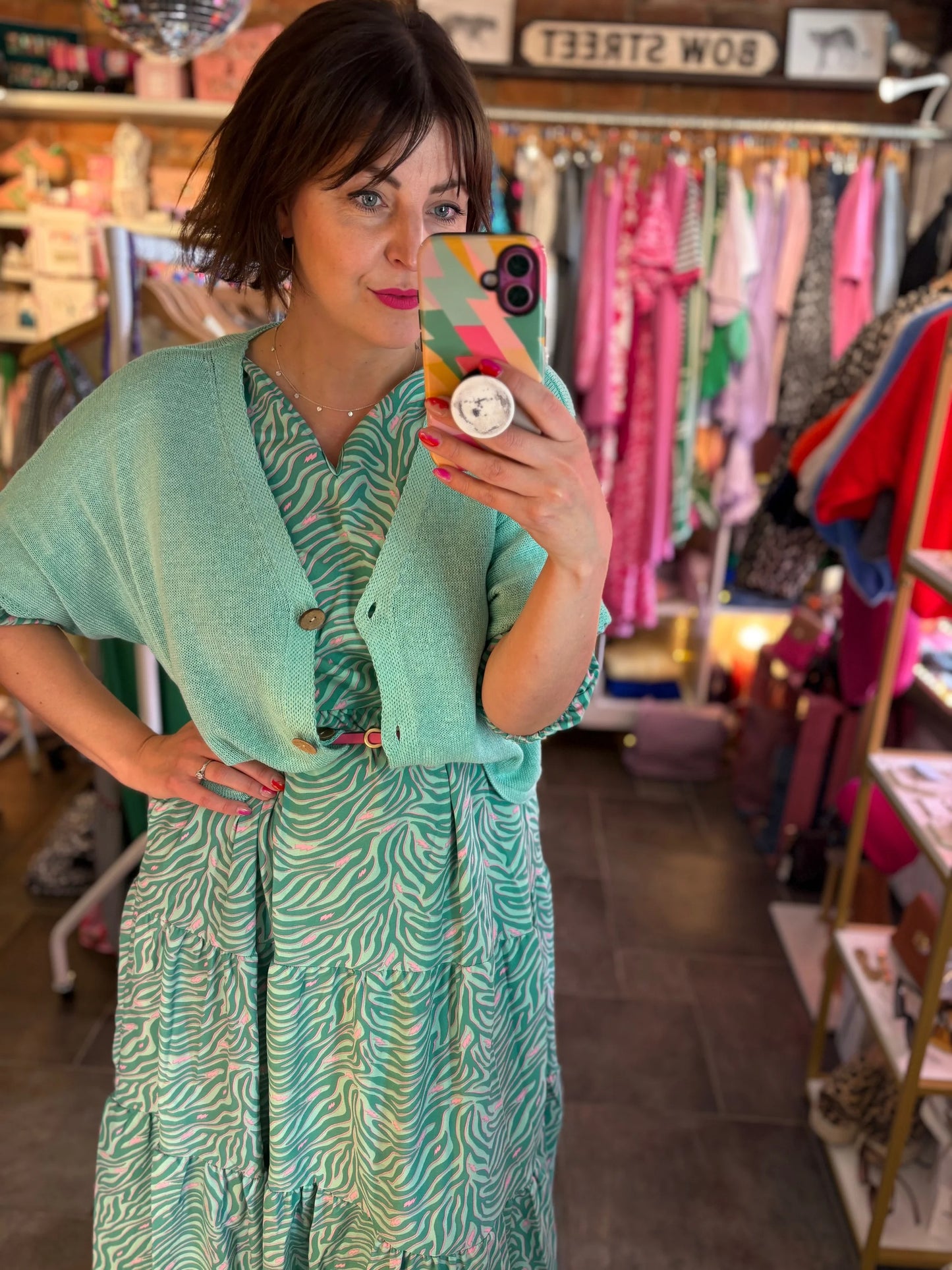 Woman taking a mirror selfie wearing a green dress with a patterned phone case in a store.