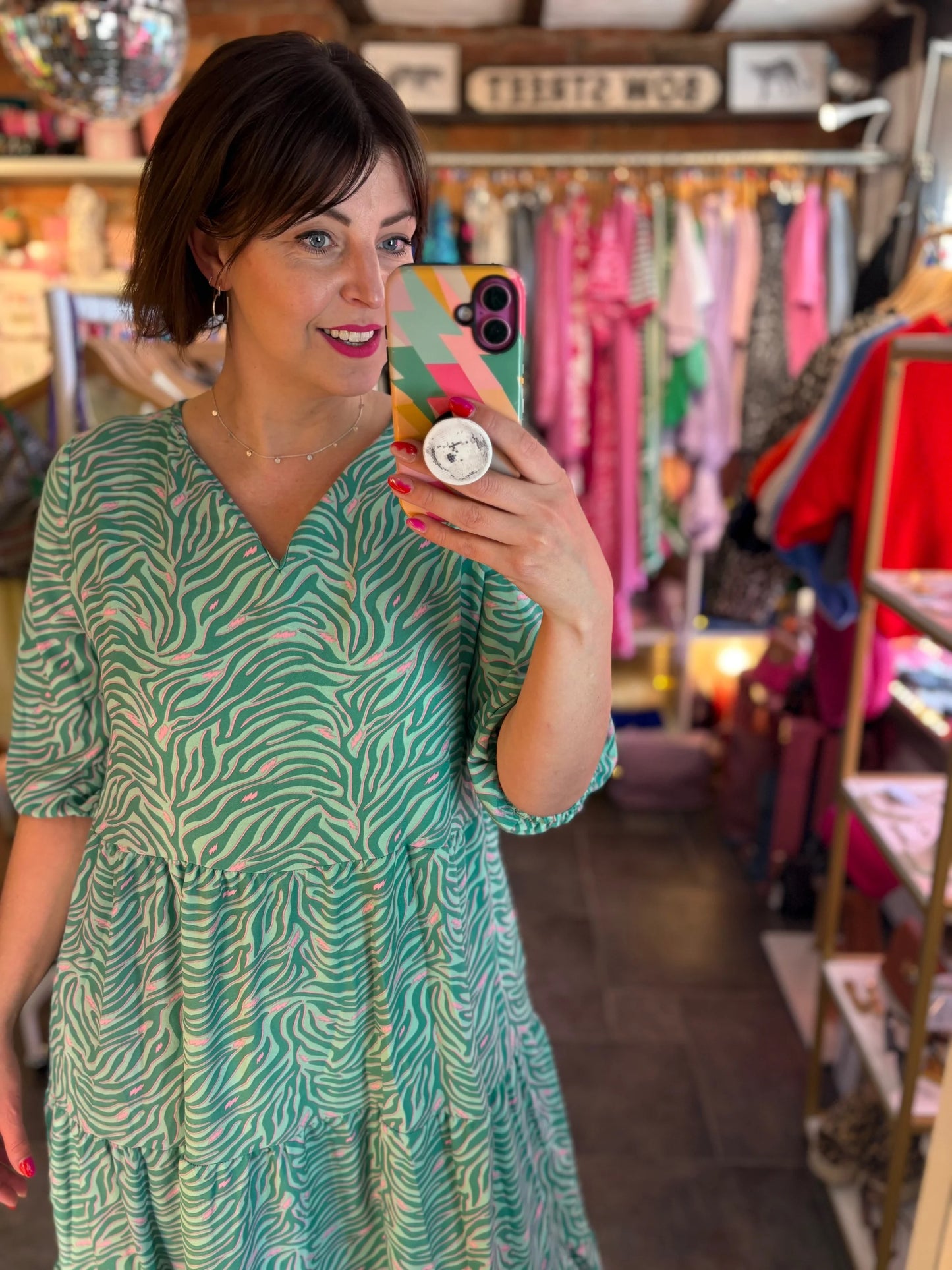 Woman taking a mirror selfie wearing a green patterned dress in a clothing store.