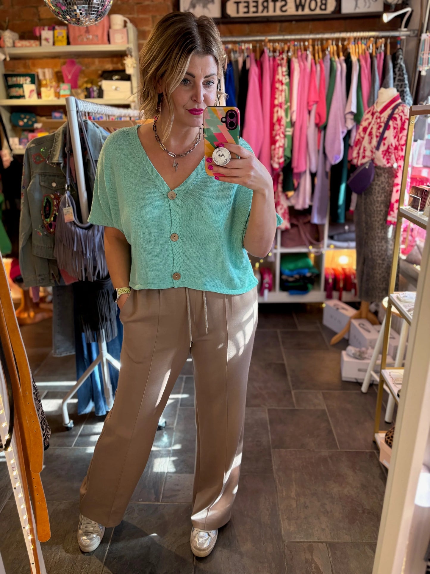 Woman taking a mirror selfie in a clothing store wearing a teal cardigan and beige pants.