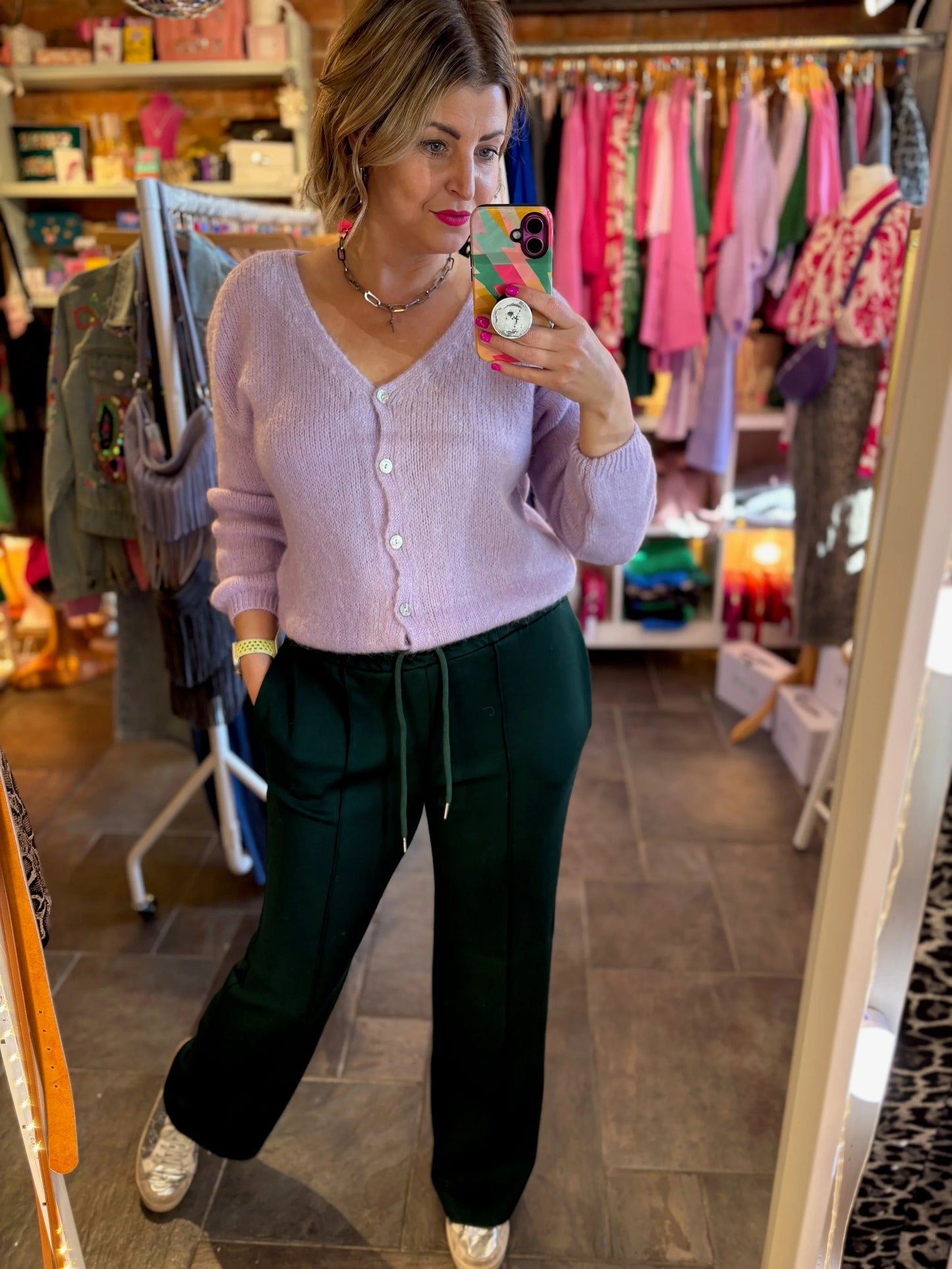 Woman taking a mirror selfie in a clothing store wearing a pink cardigan and green pants.