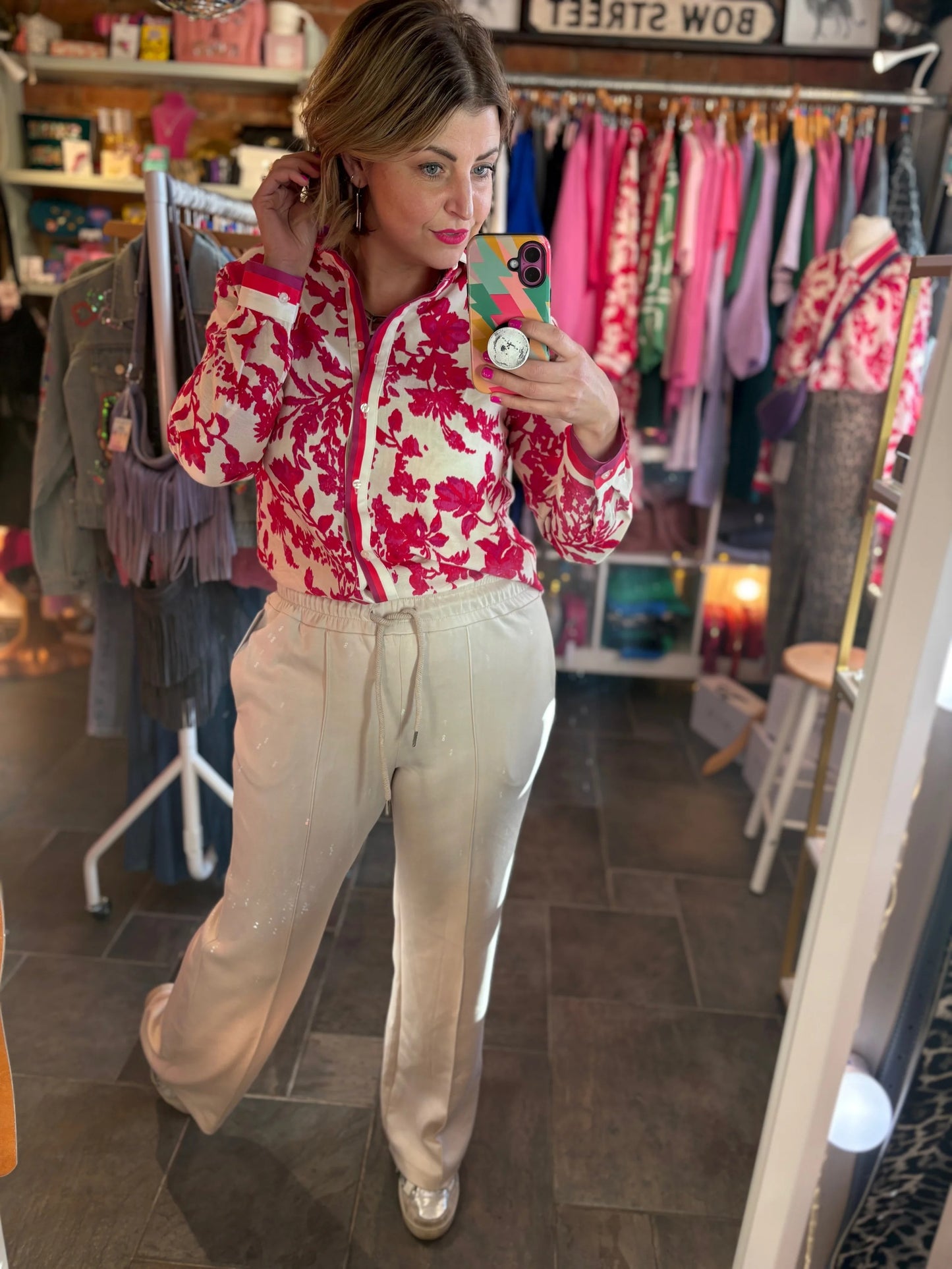 Woman taking a mirror selfie wearing a red and white floral blouse and beige pants in a clothing store.