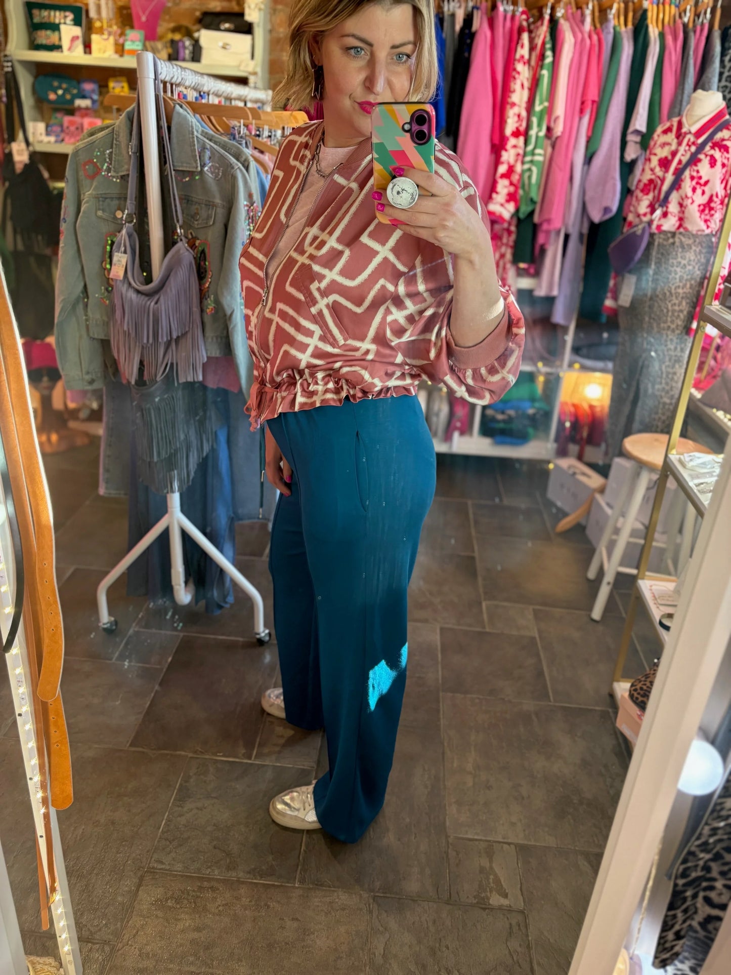 Woman taking a mirror selfie in a clothing store wearing a patterned top and blue pants.