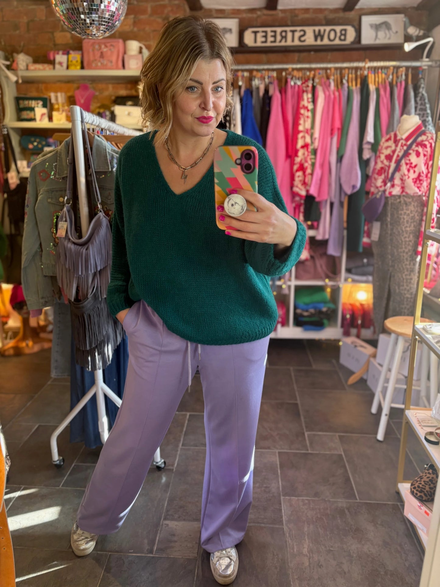 Person taking a mirror selfie wearing a green sweater and purple pants in a store.