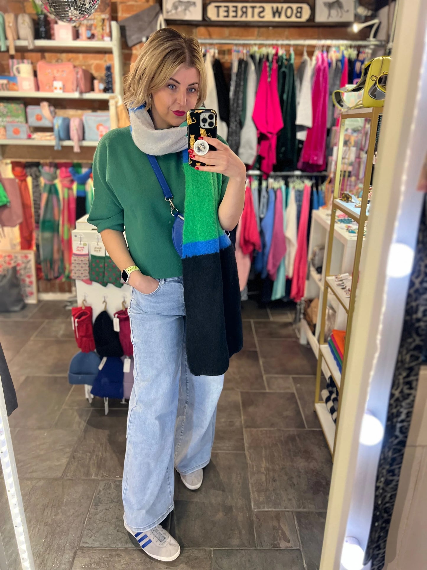 Person taking a mirror selfie in a clothing store wearing a green sweater and blue jeans.