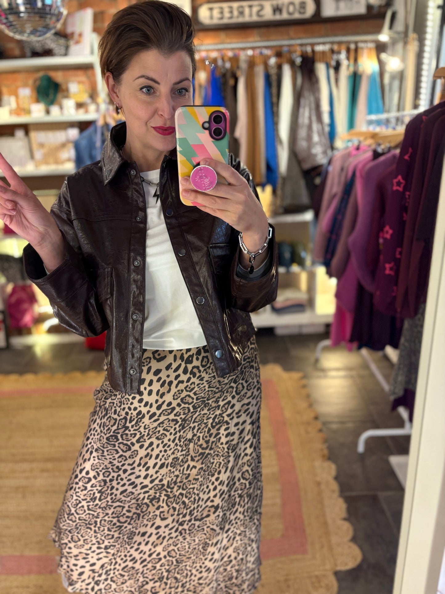 Woman taking a mirror selfie in a clothing store wearing a leopard print skirt and dark brown jacket.