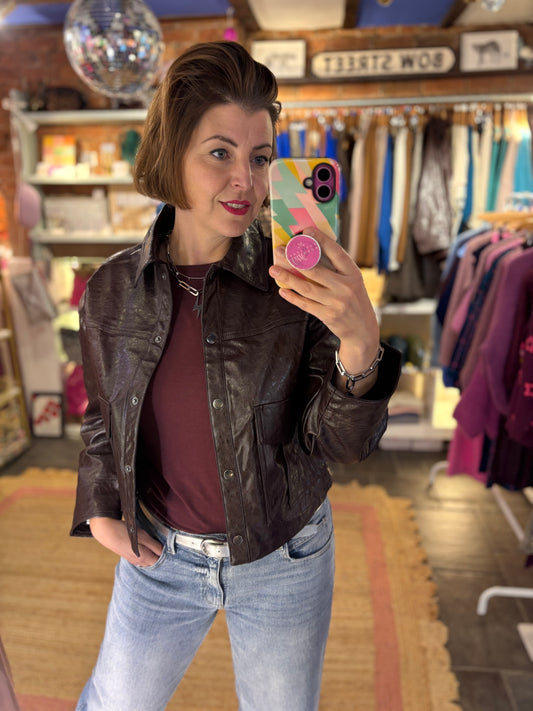 Woman taking a mirror selfie in a clothing store wearing a brown leather jacket and jeans.