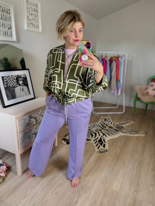 Person taking a mirror selfie wearing a green jacket and purple trousers in a room with a dresser and decor.