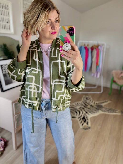 Person taking a mirror selfie wearing a khaki green patterned jacket and light blue jeans