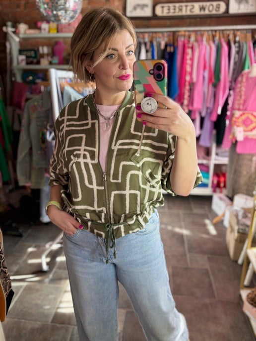 Woman taking a mirror selfie wearing a patterned khaki green jacket and jeans in a store.