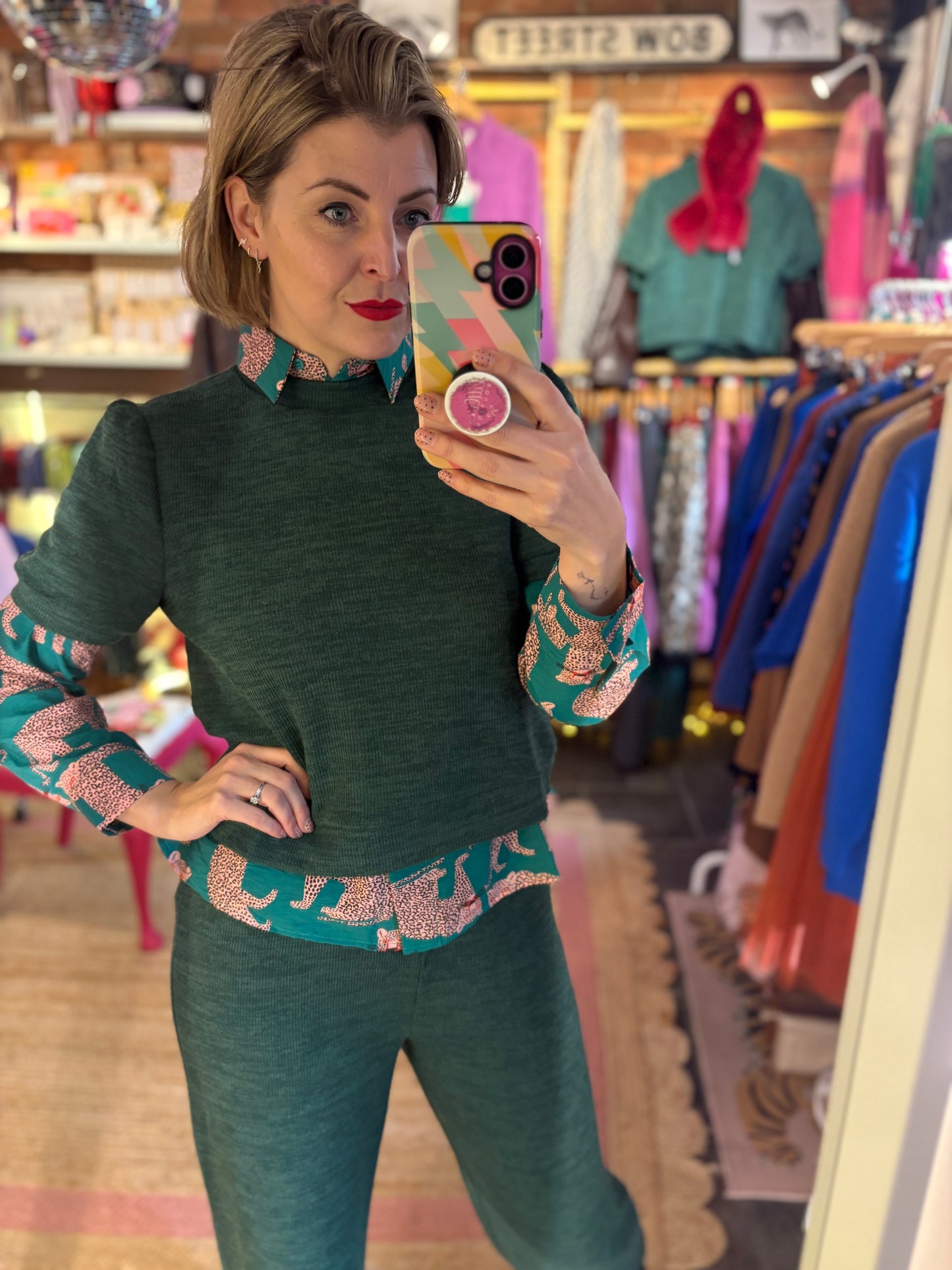 Woman taking a mirror selfie in a clothing store wearing a green co-ord outfit layered over a patterned blouse