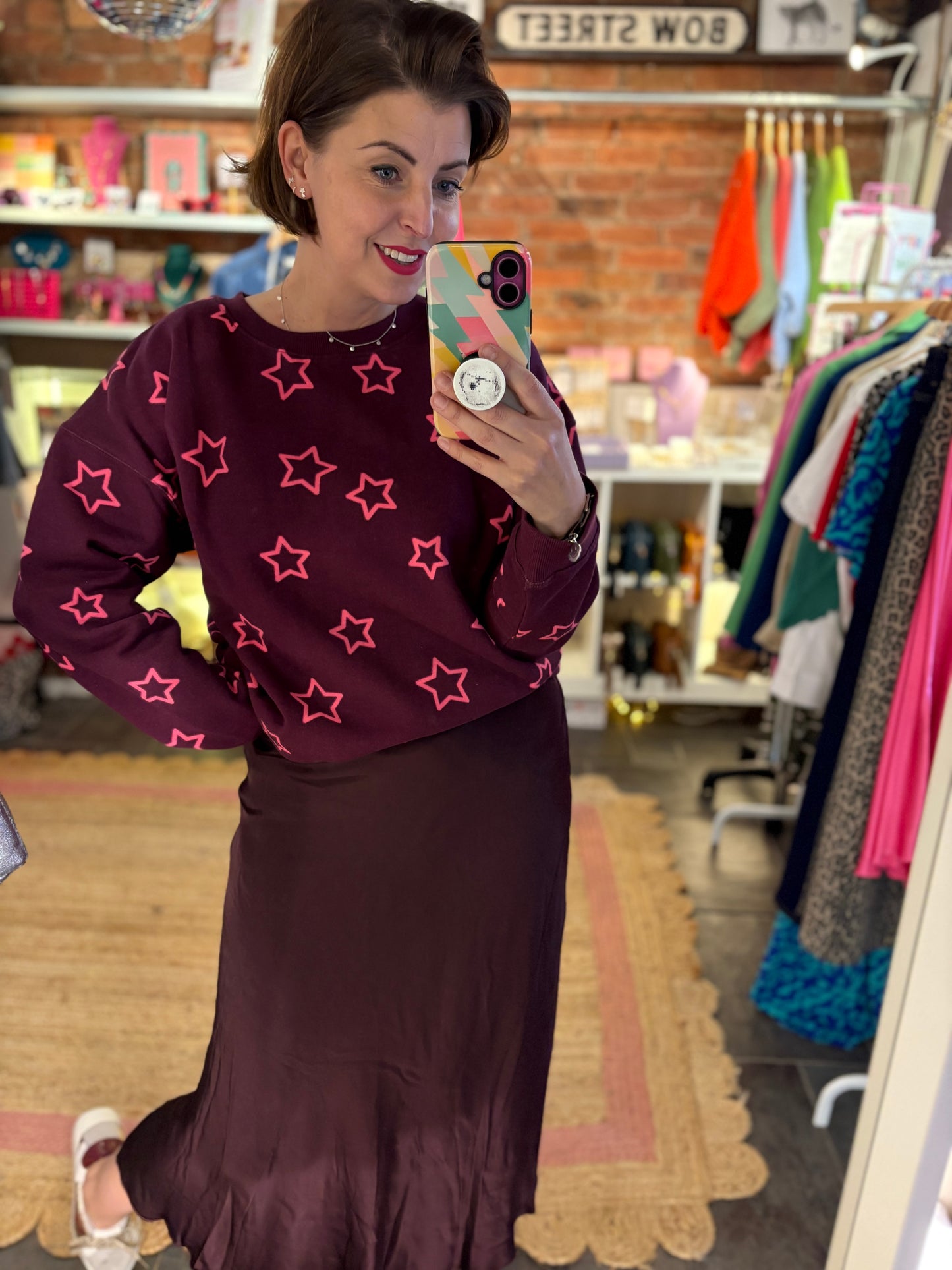 Woman taking a mirror selfie wearing a maroon sweater with pink star patterns and a matching skirt in a store.