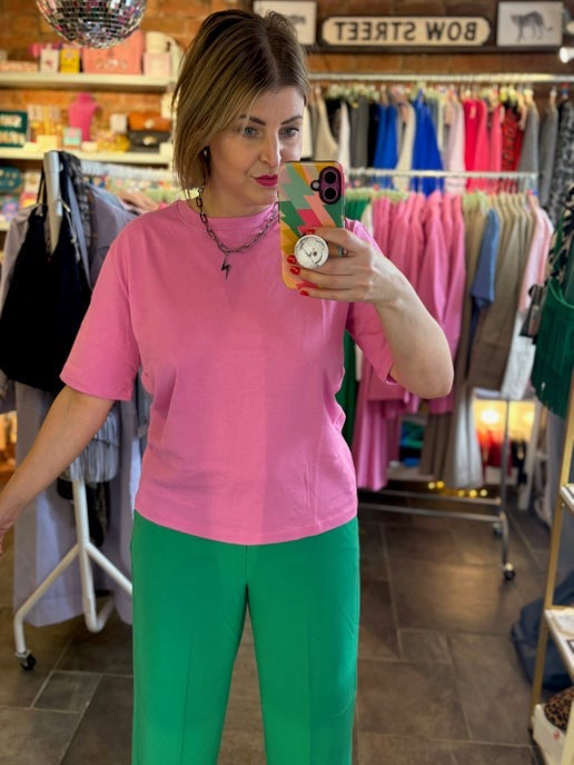 Woman taking a mirror selfie wearing a pink shirt and green pants in a clothing store.