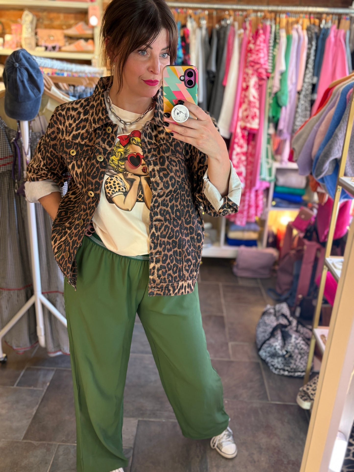 Person taking a mirror selfie wearing a leopard print jacket and green trousers in a clothing store.