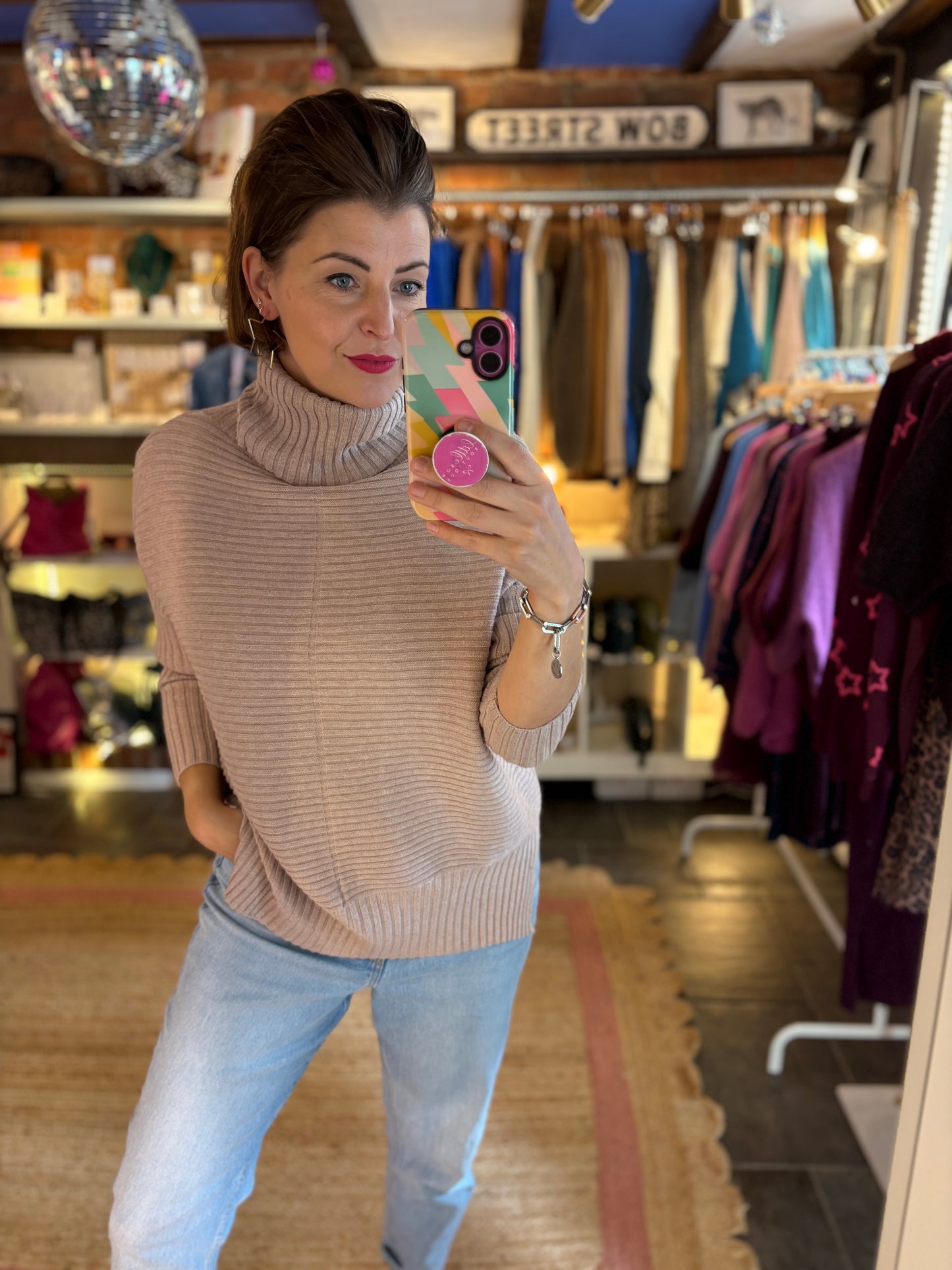 Woman taking a mirror selfie in a clothing store wearing a beige sweater and light blue jeans.