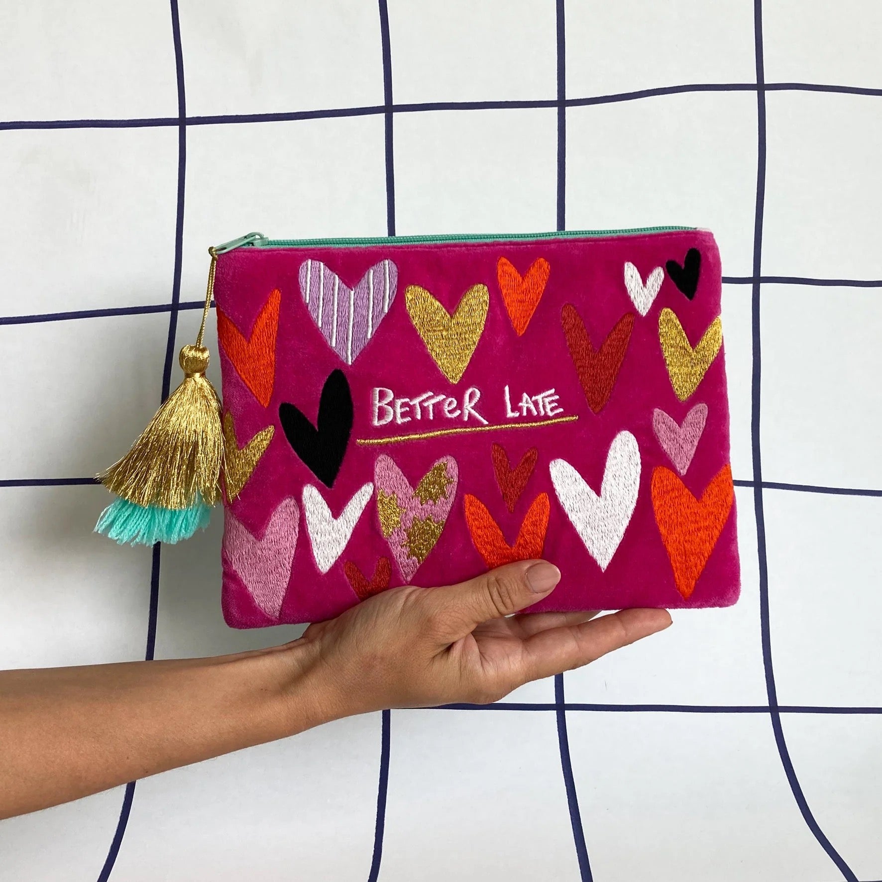 Hand holding a pink pouch with colorful heart designs and 'Better Late' text against a grid background