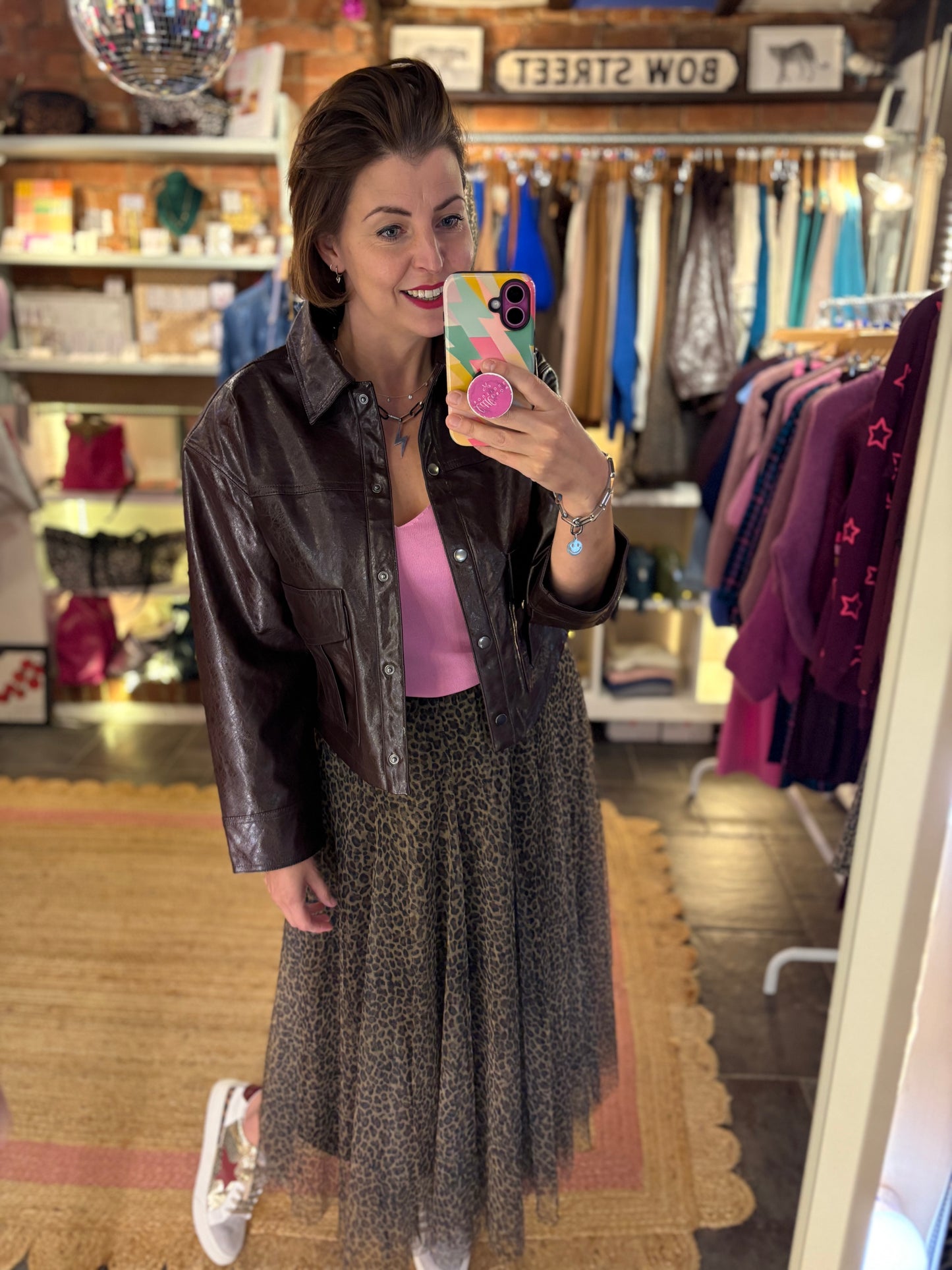 Woman taking a mirror selfie in a clothing store wearing a brown leather jacket, pink top, and leopard print patterned tulle skirt.