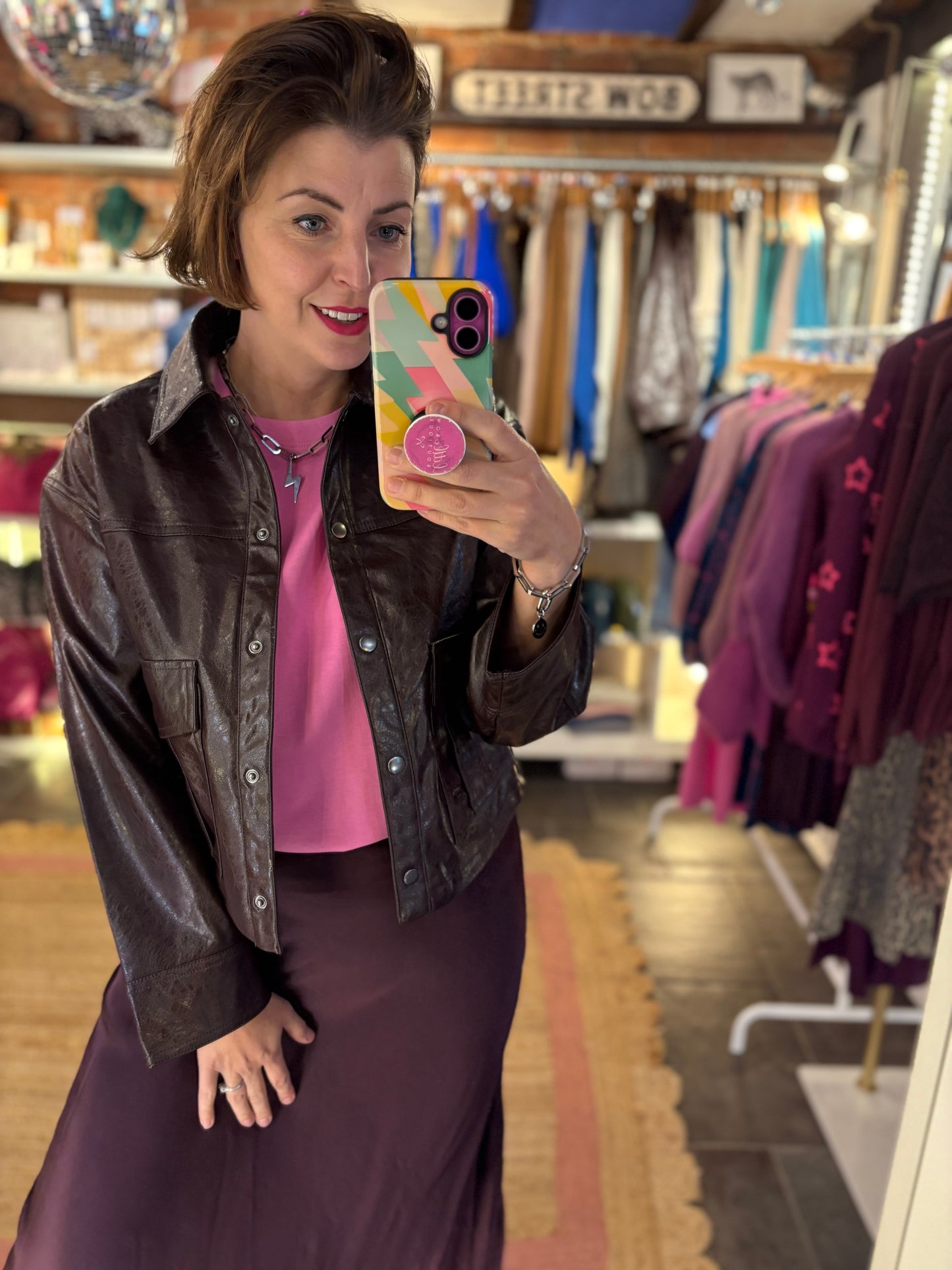 Woman taking a mirror selfie in a clothing store wearing a brown leather jacket and pink shirt.