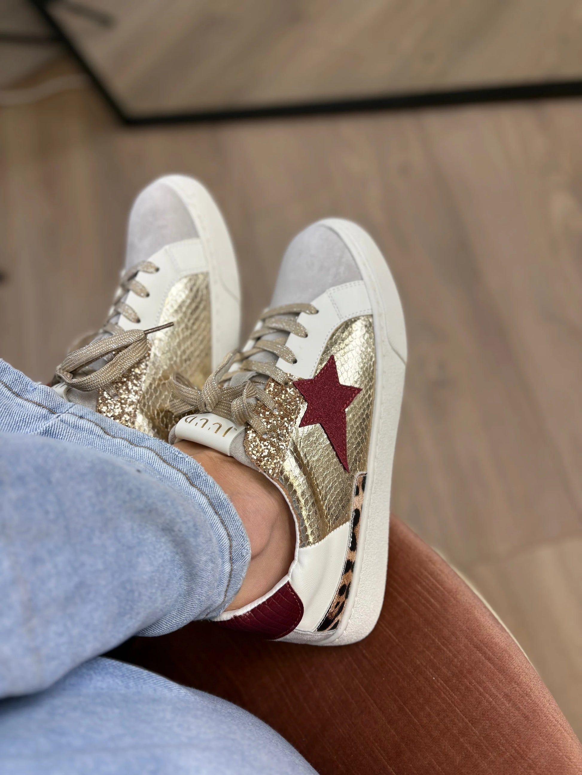 White sneakers with gold accents and a red star worn by a person sitting on a wooden floor.