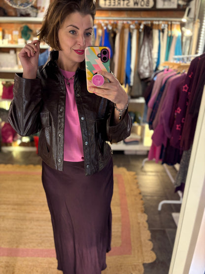 Woman taking a mirror selfie in a clothing store wearing a brown jacket and pink shirt.