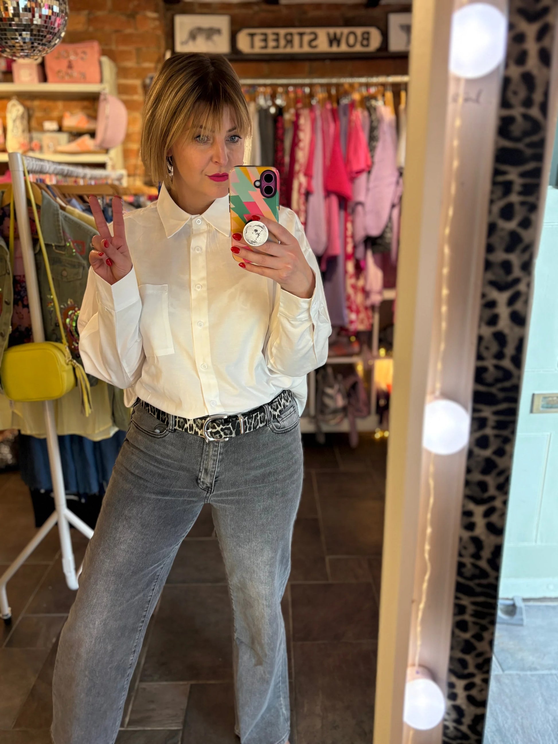 Person taking a mirror selfie wearing a white shirt and gray jeans in a store.