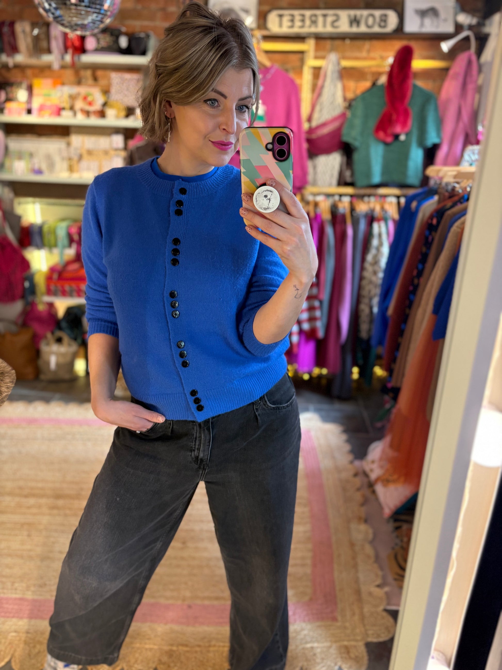 Woman taking a mirror selfie wearing a blue cardigan and black jeans in a store.